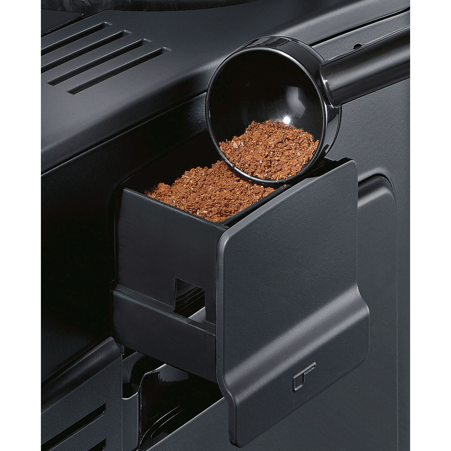 Coffee grounds being scooped into a coffee machine compartment.