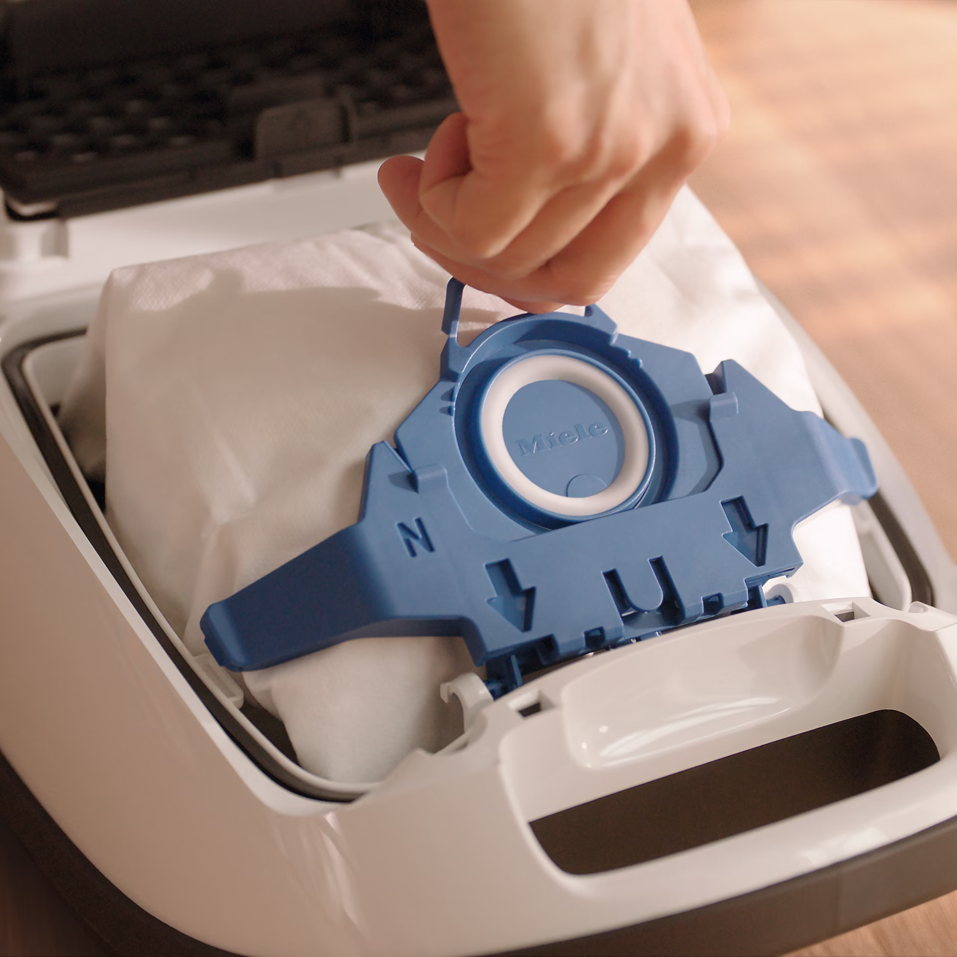 Dłoń trzymająca niebieski uchwyt worka do odkurzacza. Biała torba wewnątrz białego odkurzacza.