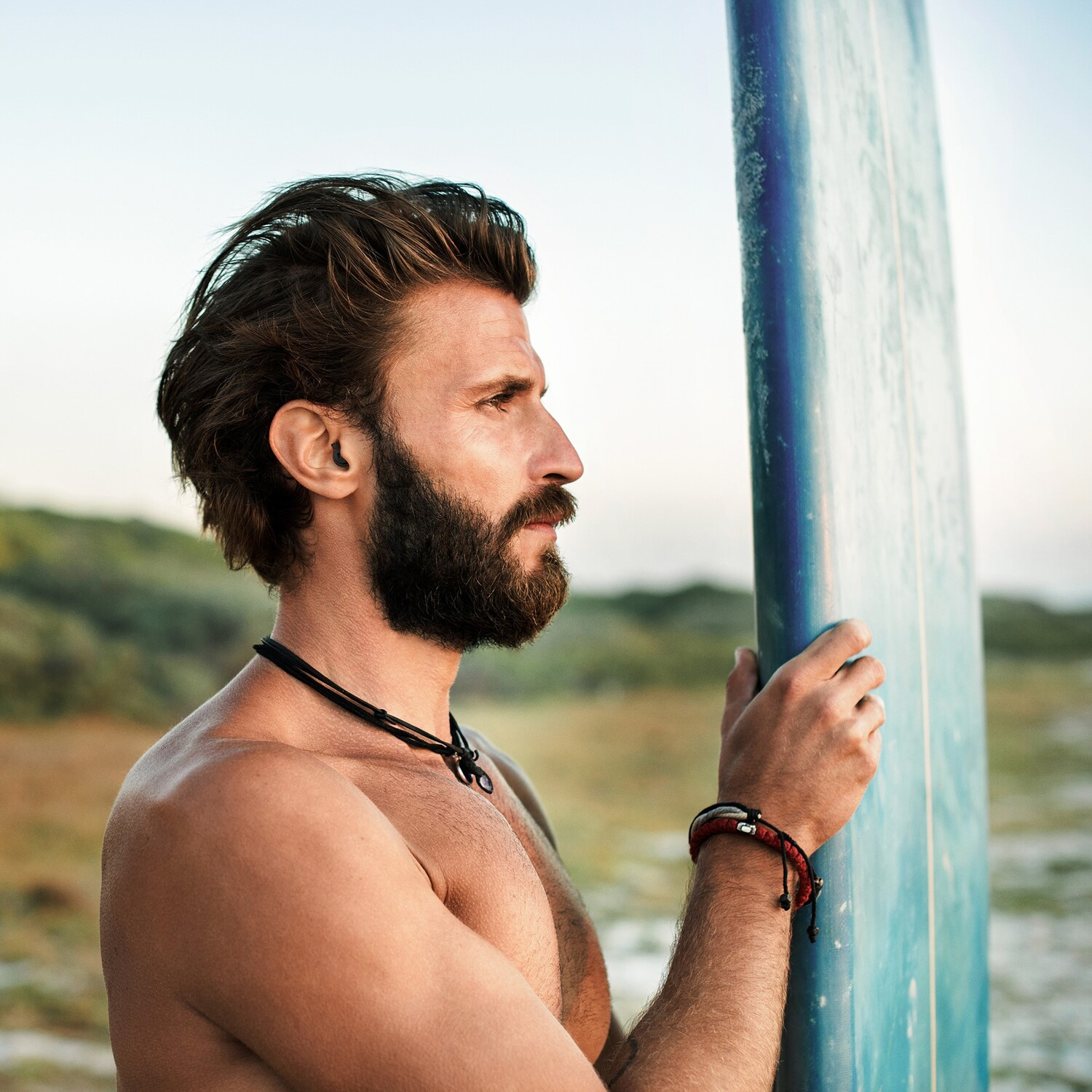 Man with beard holding a blue surfboard, looking towards the sea. He is shirtless.