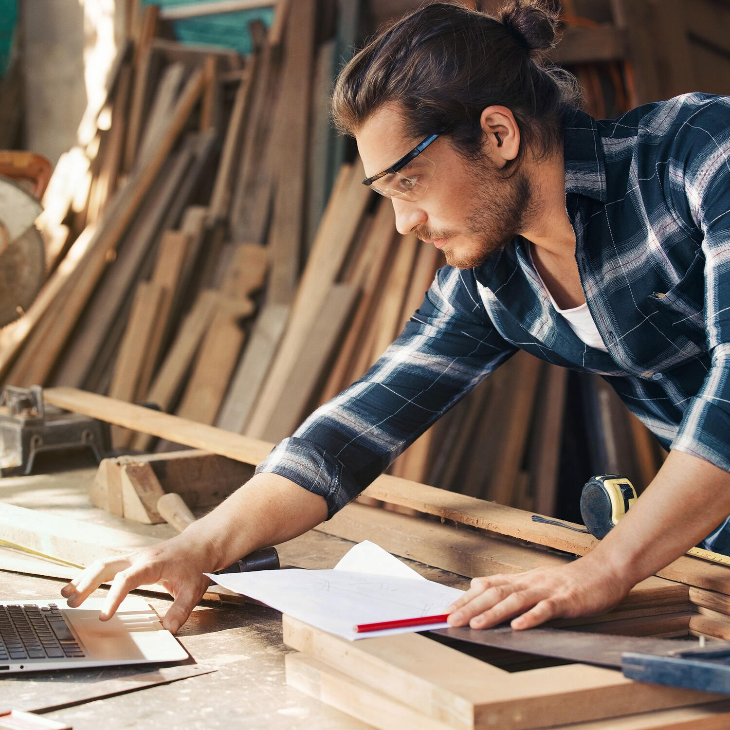 Homme travaillant sur un ordinateur portable dans un atelier, entouré de bois et d'outils.
