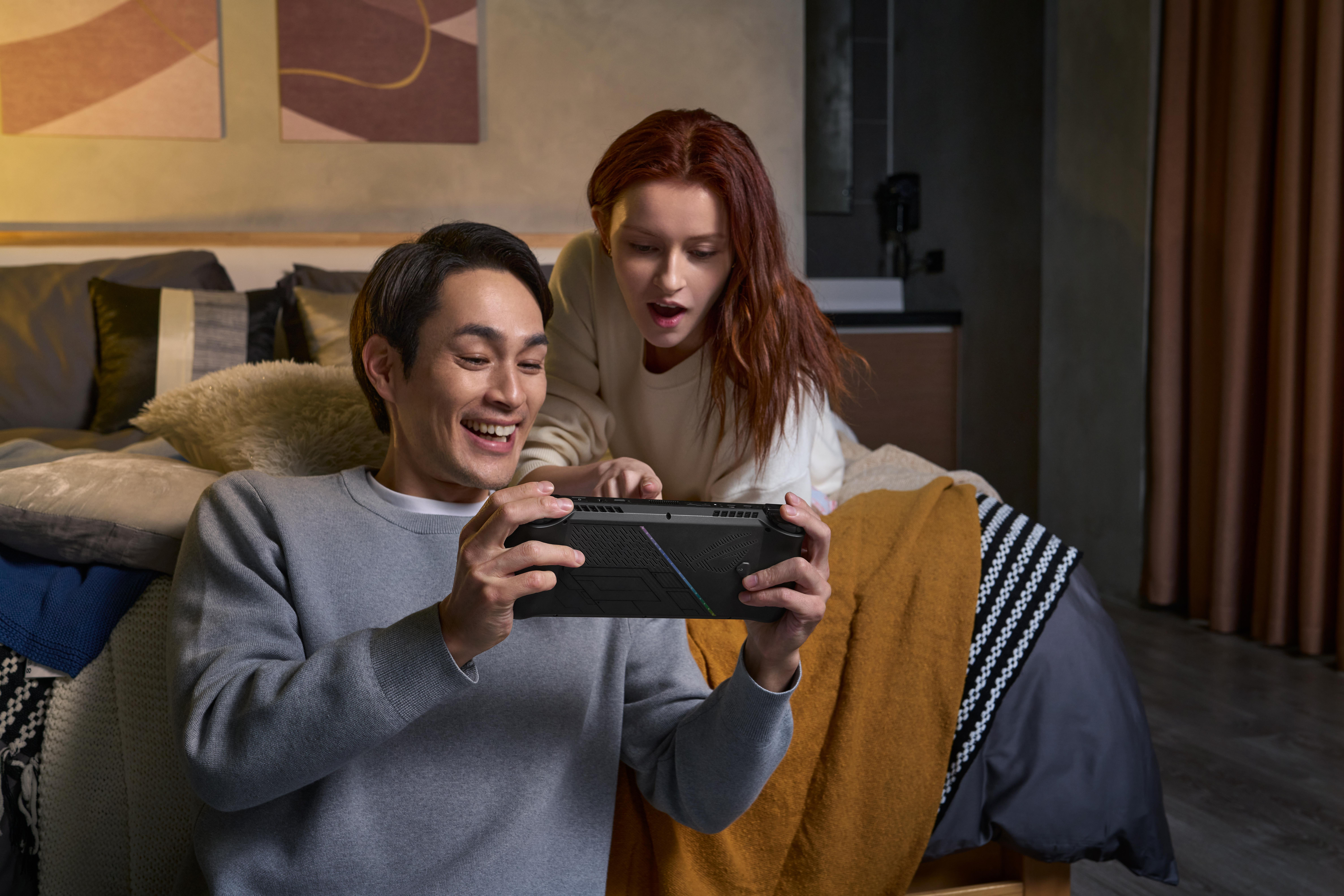 Ein Paar spielt gemeinsam ein Handheld-Spiel, lächelnd. Bett und Kissen im Hintergrund, warmes Licht.