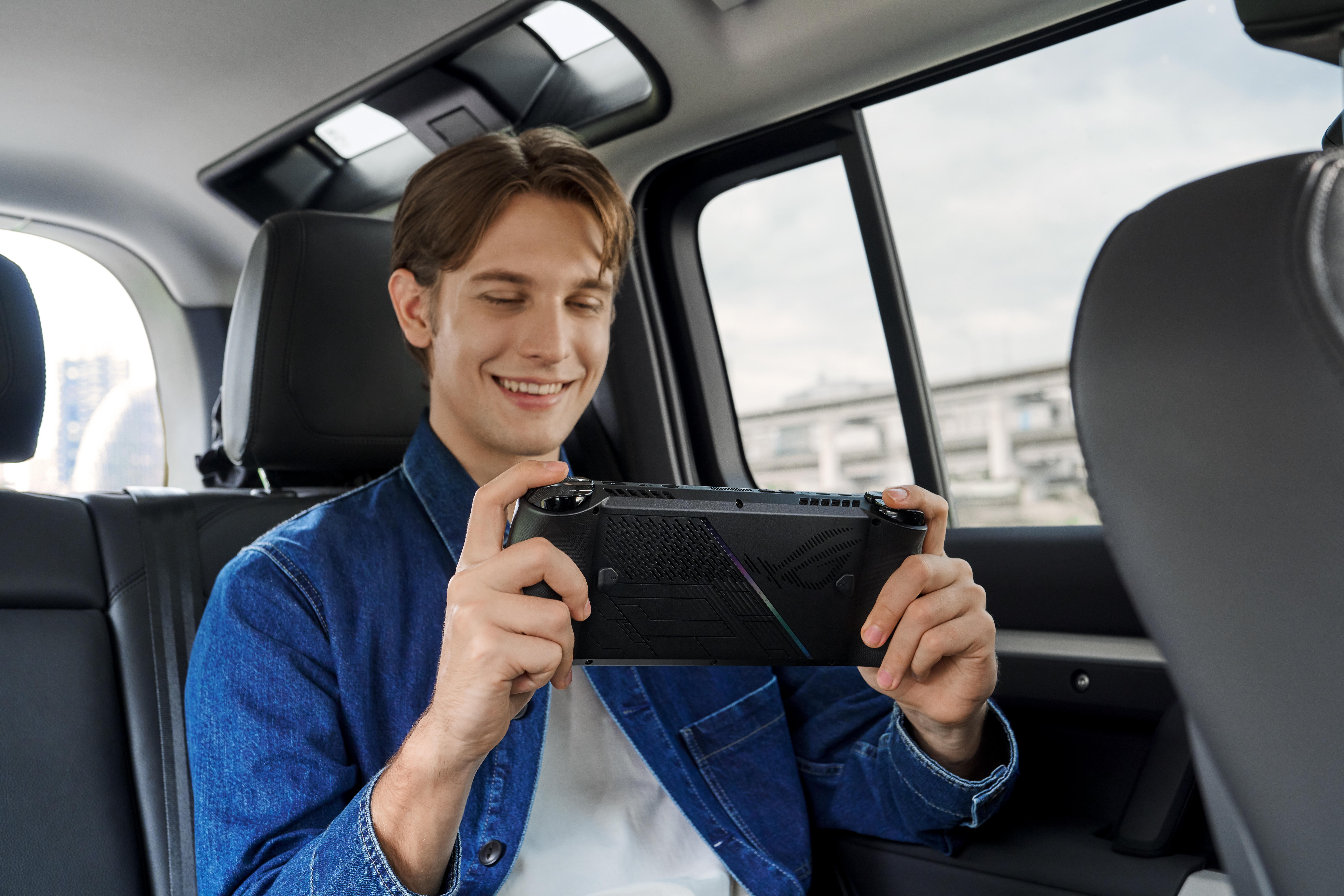 Mann lächelt, hält ein Gaming-Gerät. Sitzt in einem Auto. Dunkles Interieur.