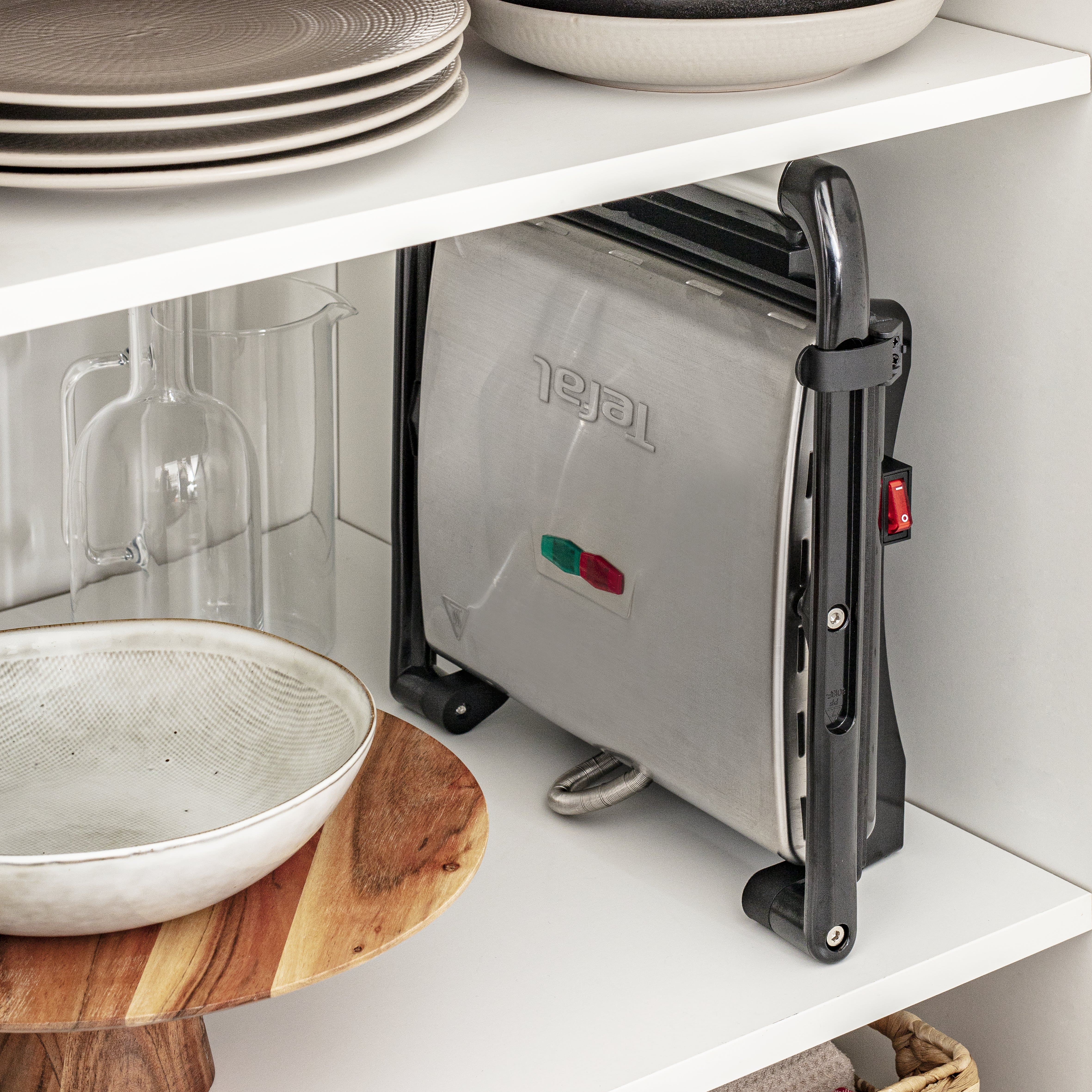 Parrilla Tefal dentro de armario blanco. Platos, vidrio y tabla de madera presentes.