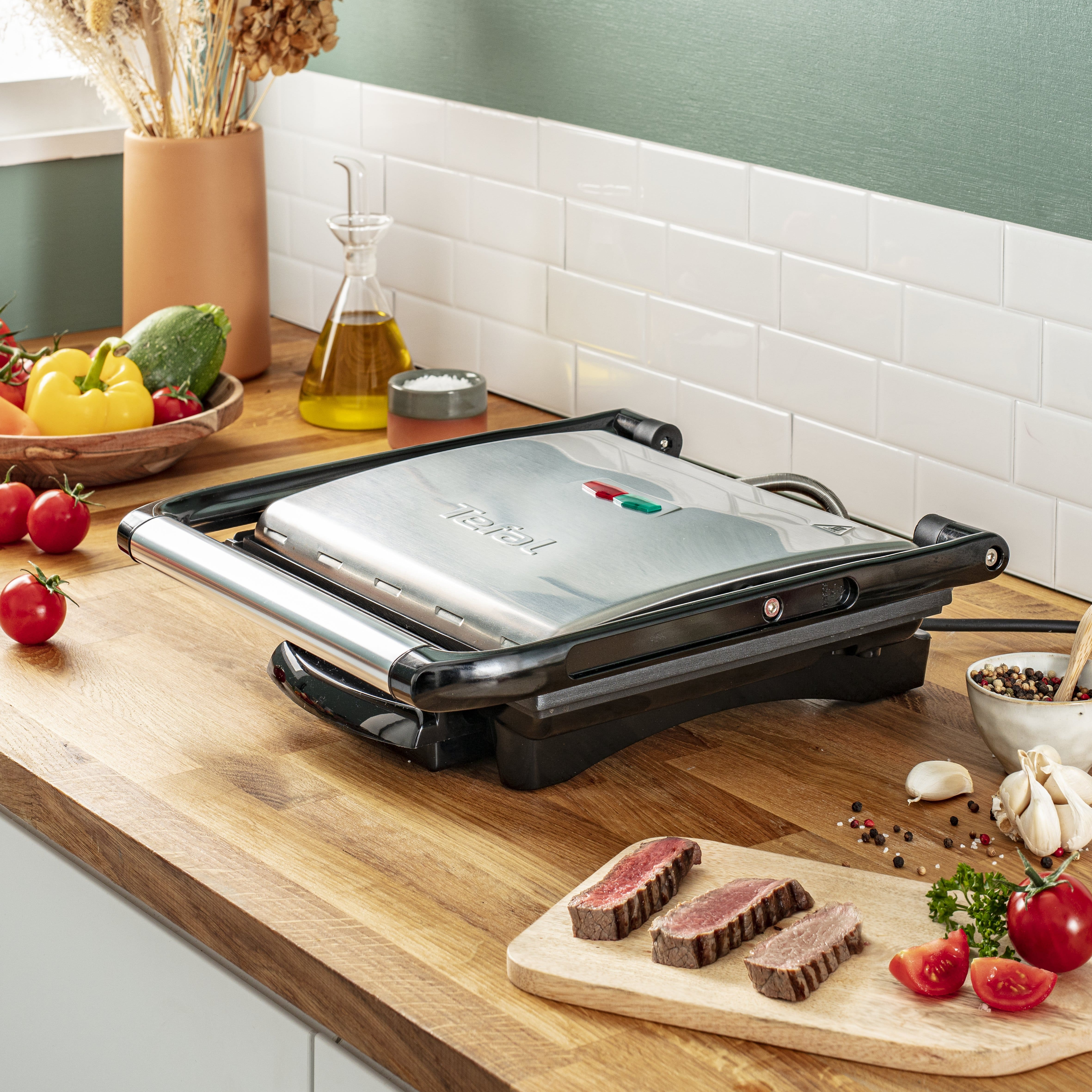 Una parrilla Tefal plateada sobre encimera de madera con tomates, carne, verduras y hierbas.