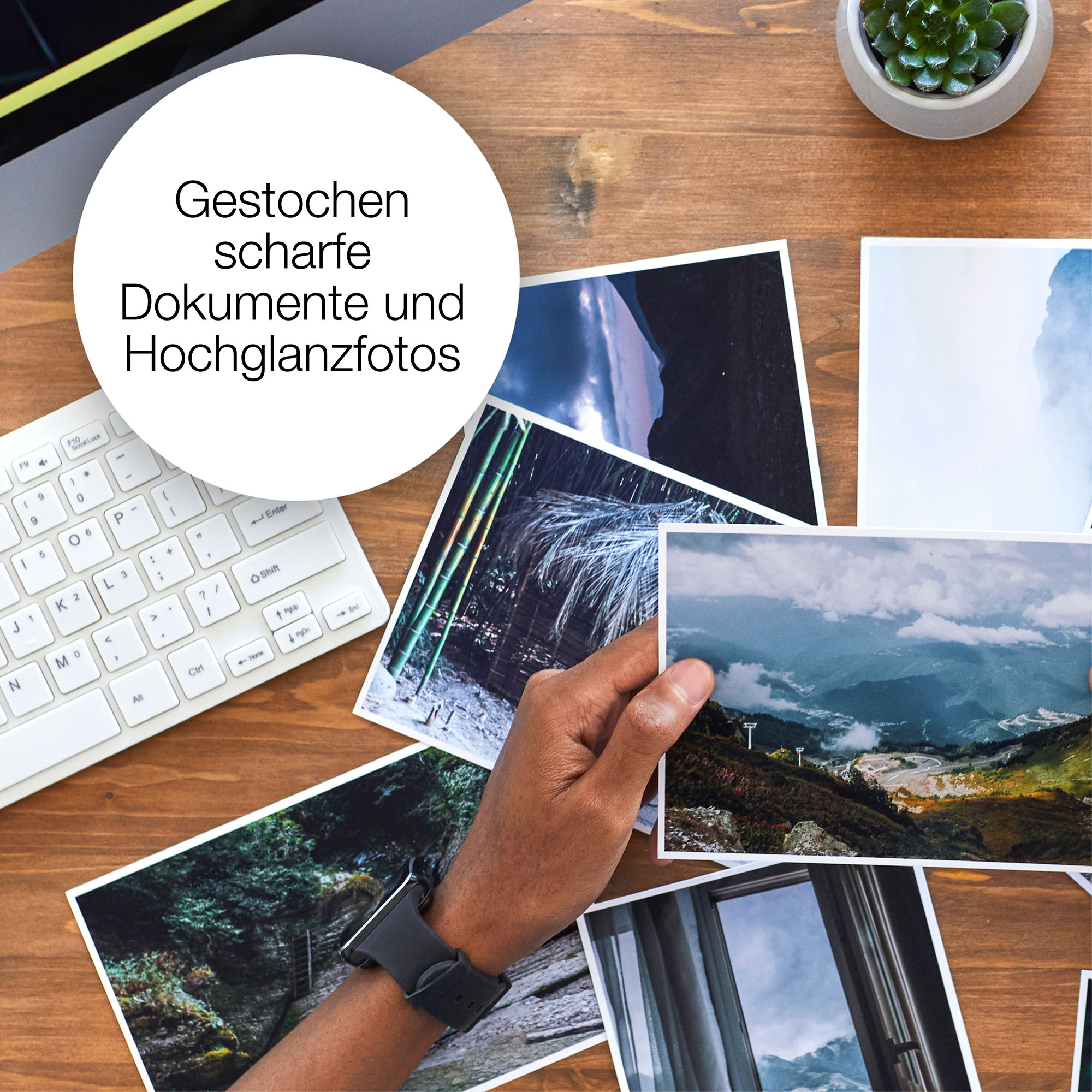 Fotos auf einem Holztisch, eine Hand hält eine. Tastatur und Pflanze im Hintergrund.