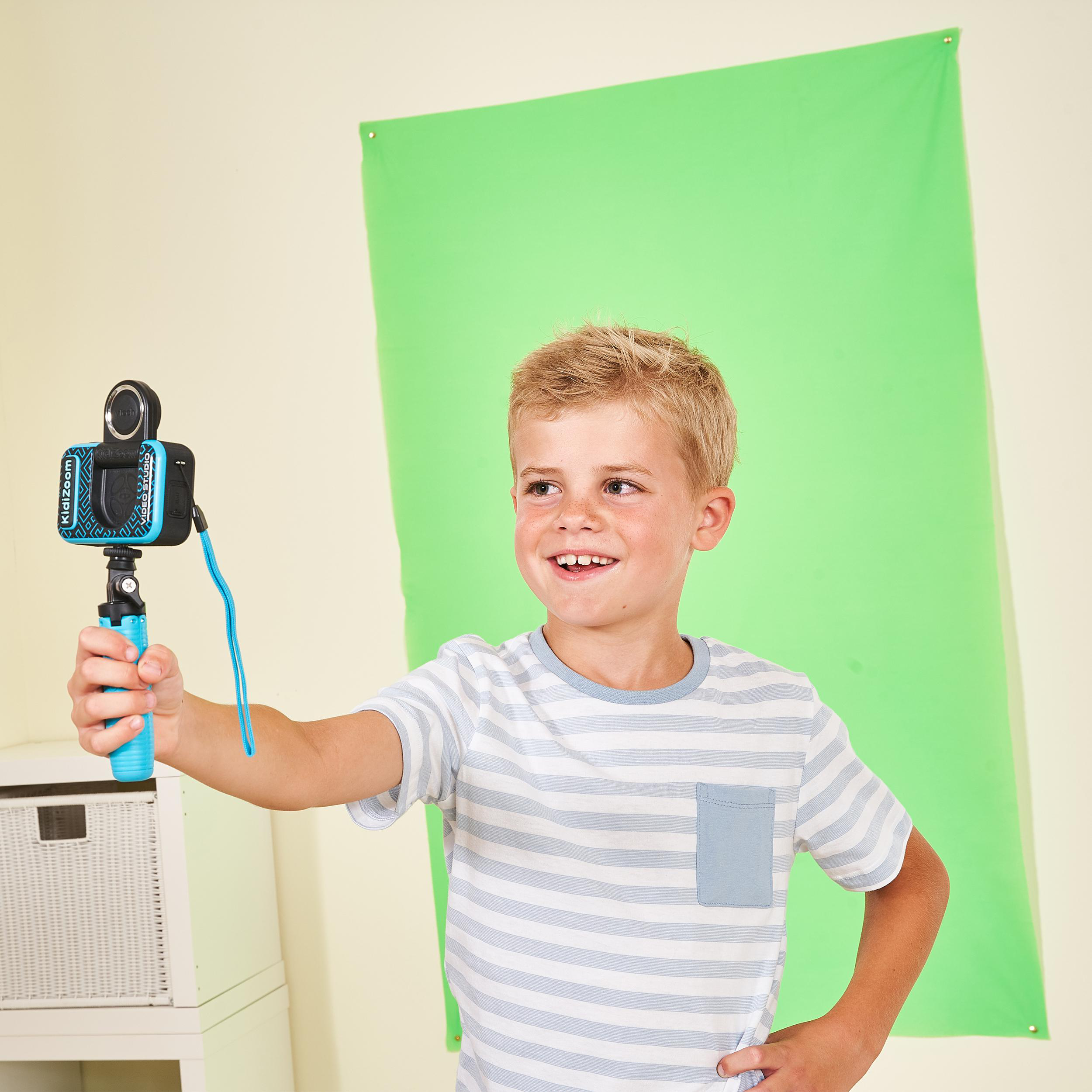 Ein Junge hält eine Kamera auf einem Selfie-Stick und lächelt vor einem grünen Hintergrund.
