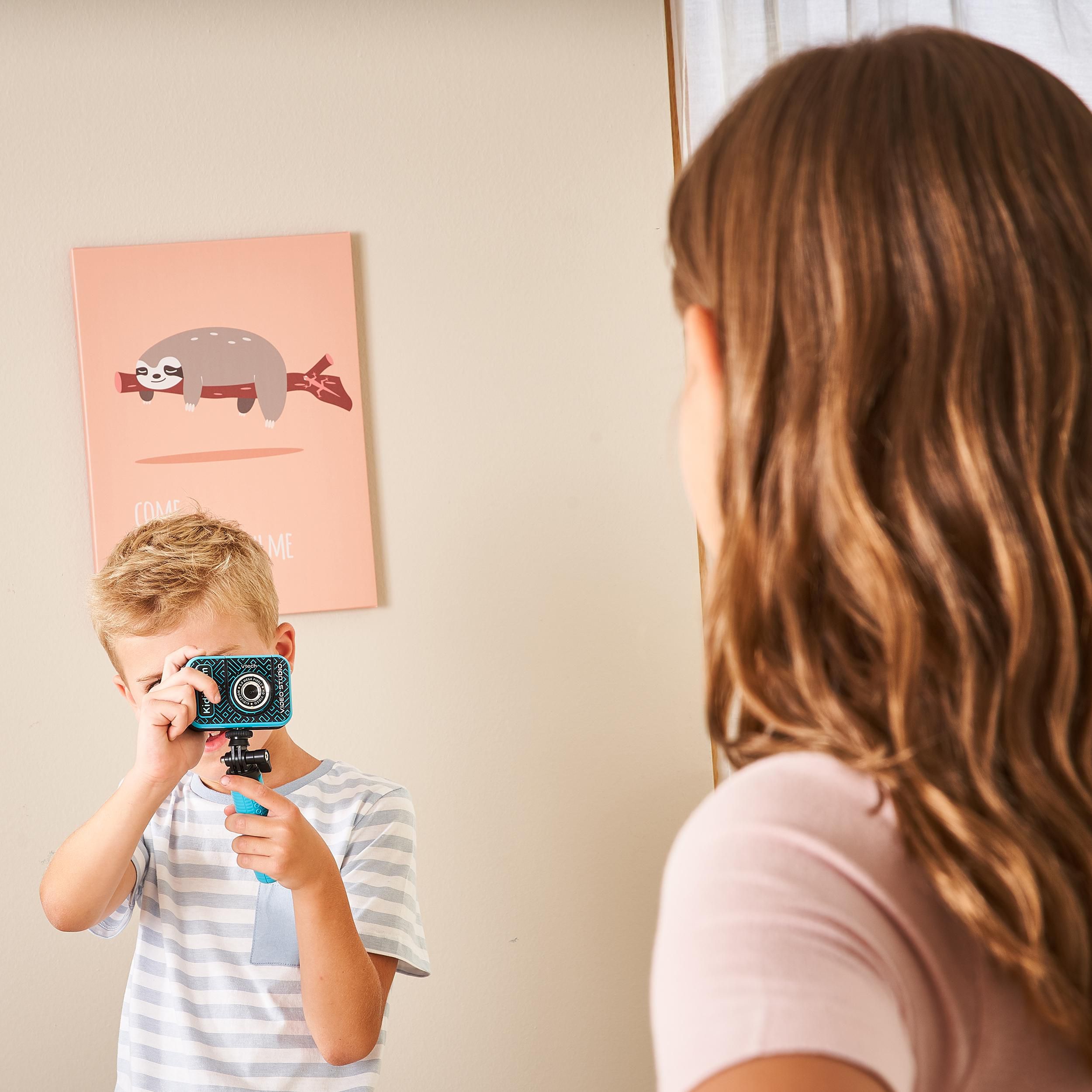 Ein Junge macht mit einer blauen Kamera ein Foto einer Frau. Ein Faultierbild hängt an der Wand.