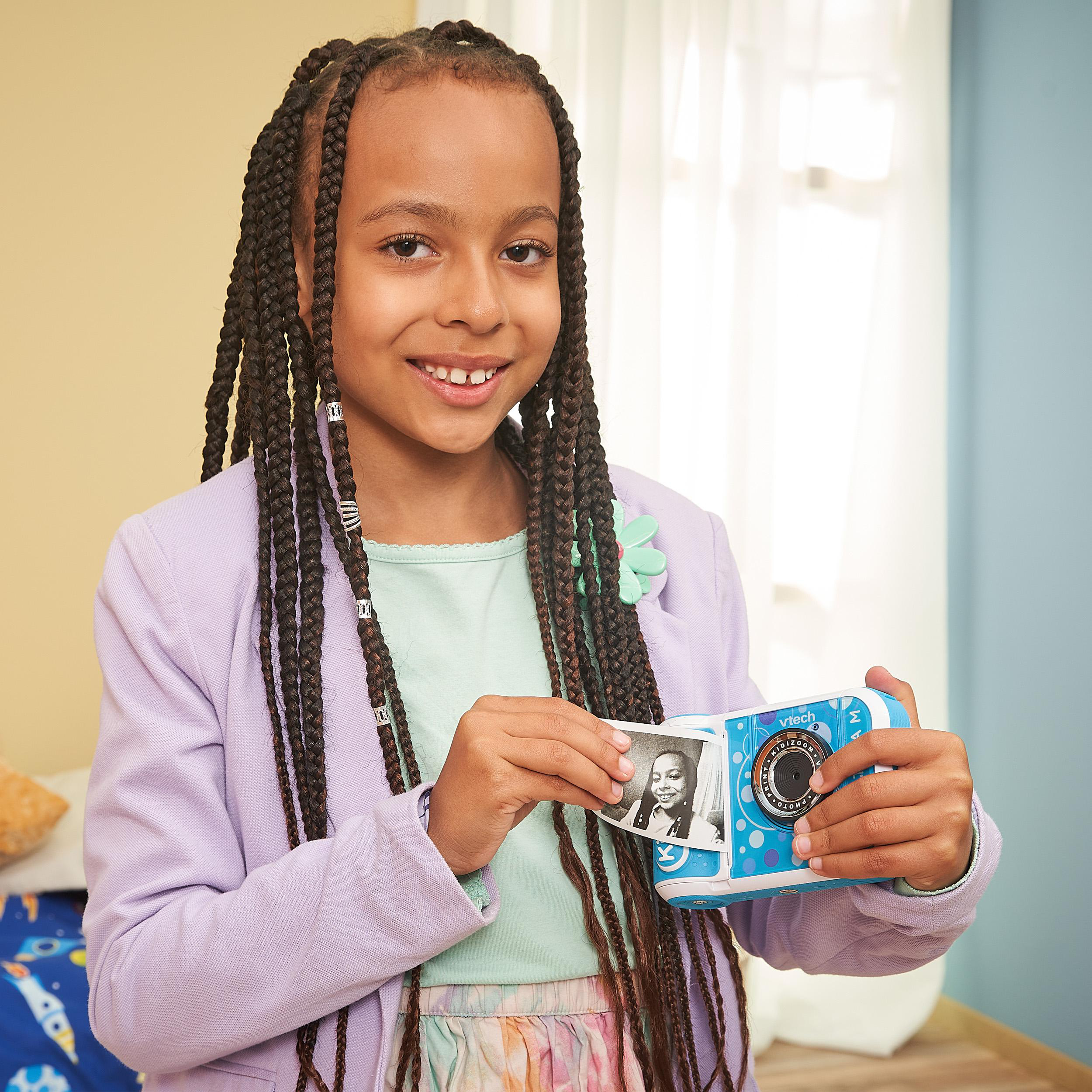 Junges Mädchen mit Zöpfen hält Spielzeugkamera, lächelt, blaue Kamera, helllila Jacke.