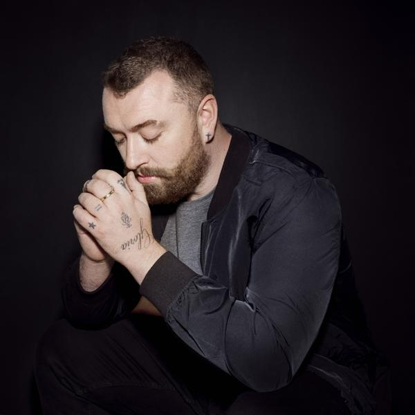 Sam Smith kniet mit gefalteten Händen, Tattoos sichtbar. Er trägt eine Bomberjacke und dunkle Kleidung.
