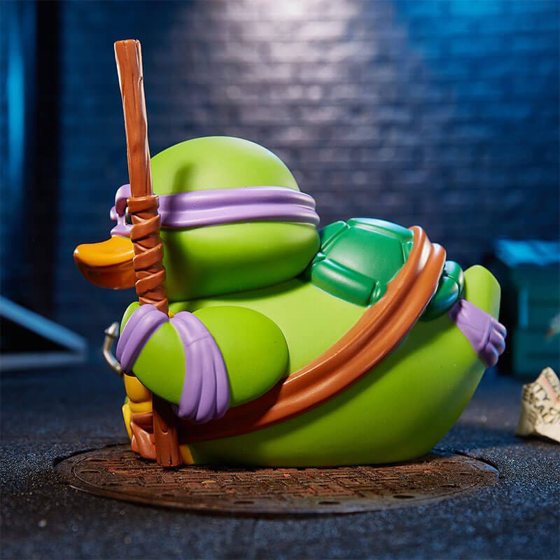 Egy Donatello gumikacsa játék, zöld páncéllal és lila maszkkal, egy csatornában.