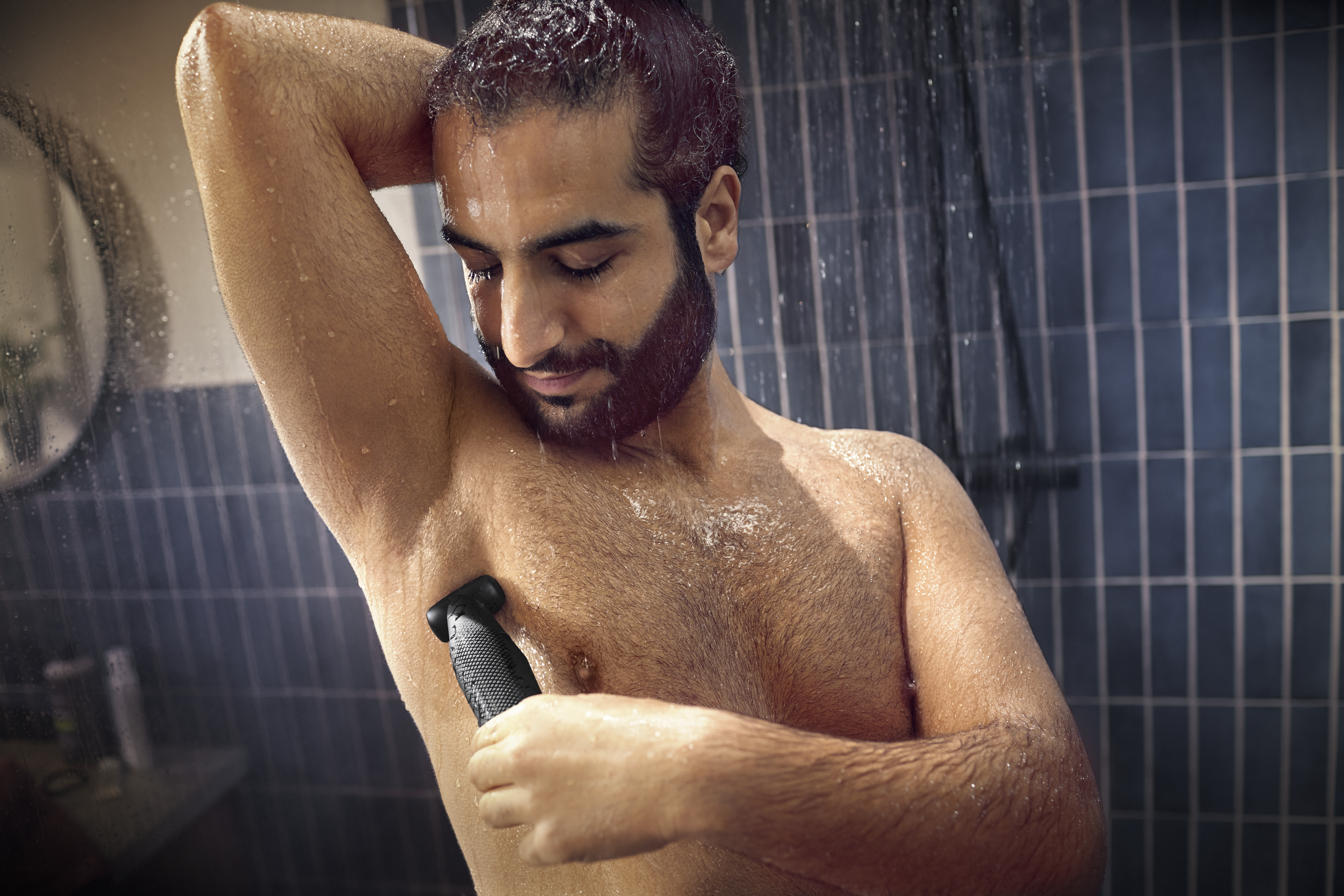 Hombre afeitándose el vello de las axilas con una recortadora en la ducha.