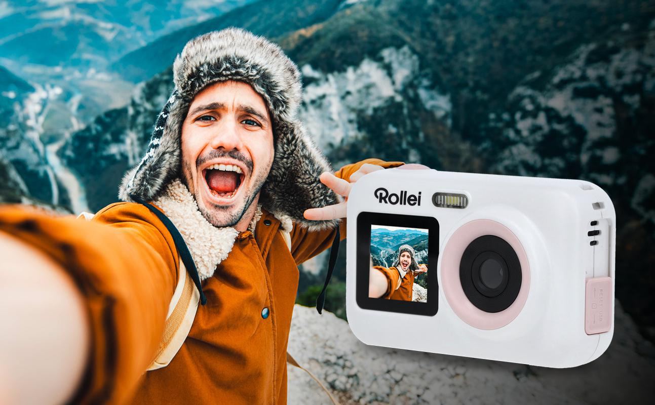 Ein Mann macht ein Selfie mit einer weißen Kamera auf einem Berg, lächelnd.