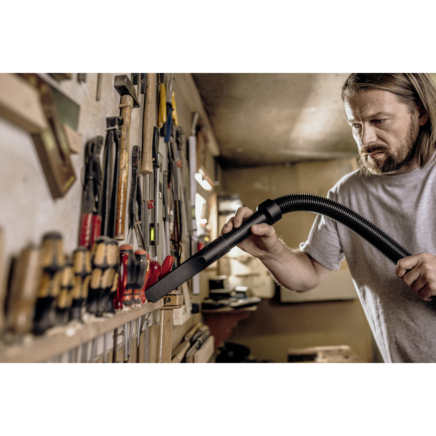 A man using a vacuum cleaner in a workshop with tools on a wall.