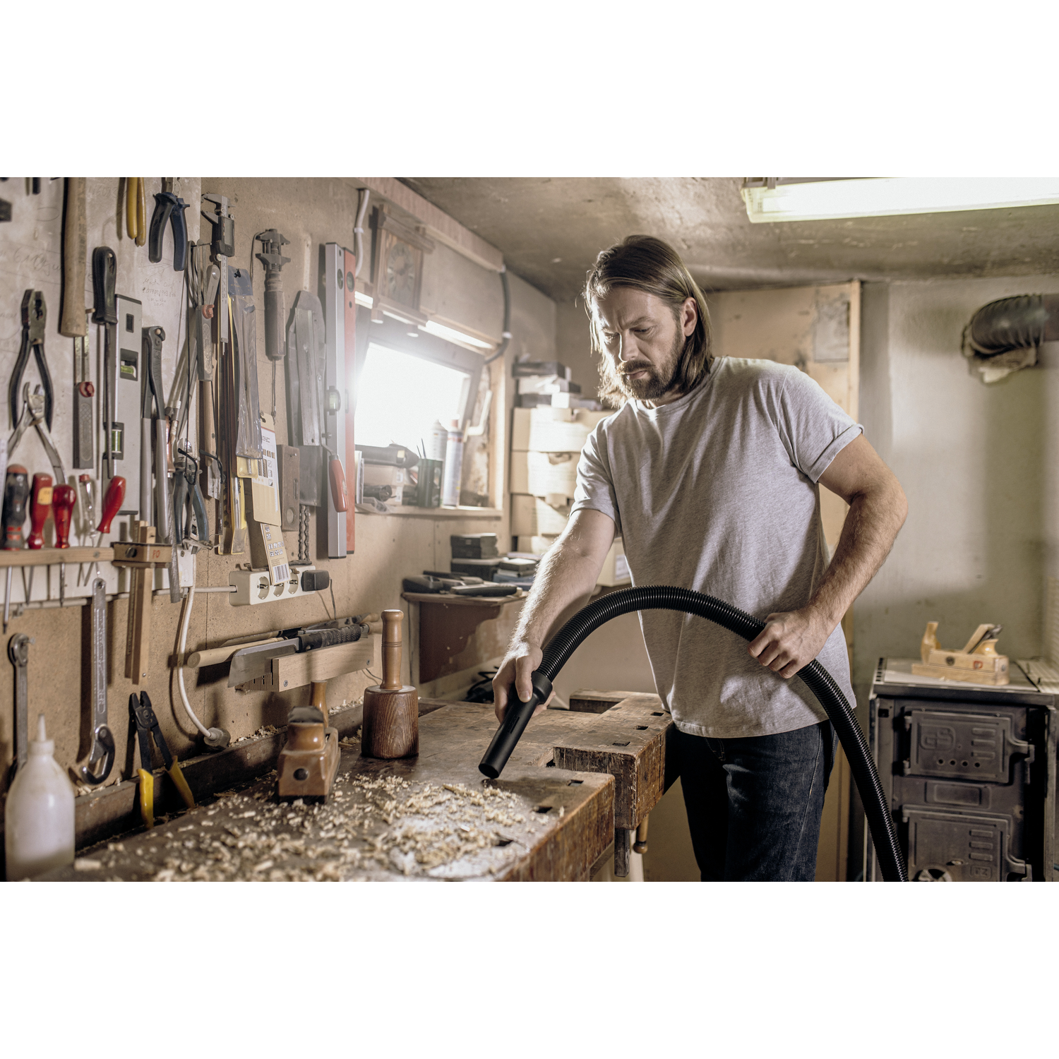 A man in a workshop uses a vacuum cleaner on a workbench covered in wood shavings.