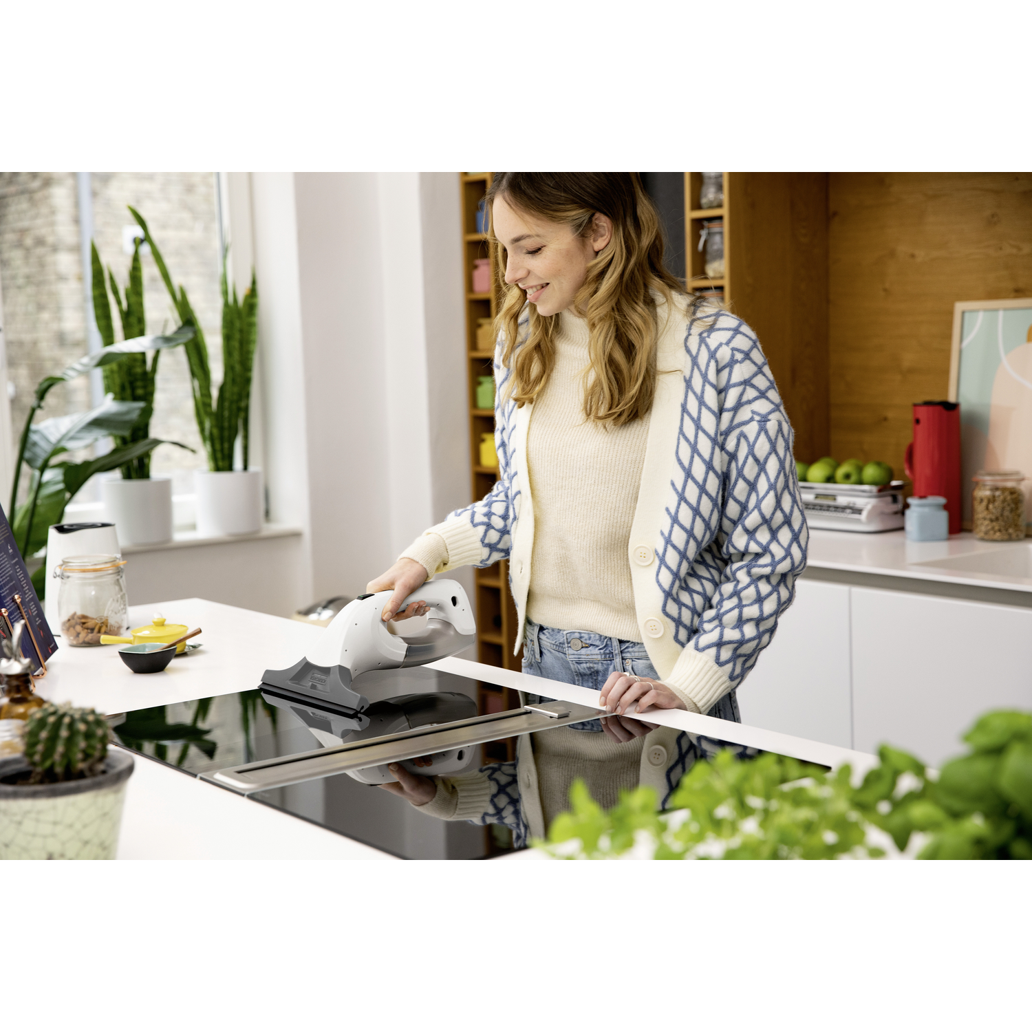 Une femme nettoyant une table de cuisson noire avec un outil de nettoyage, dans un environnement de cuisine lumineux.