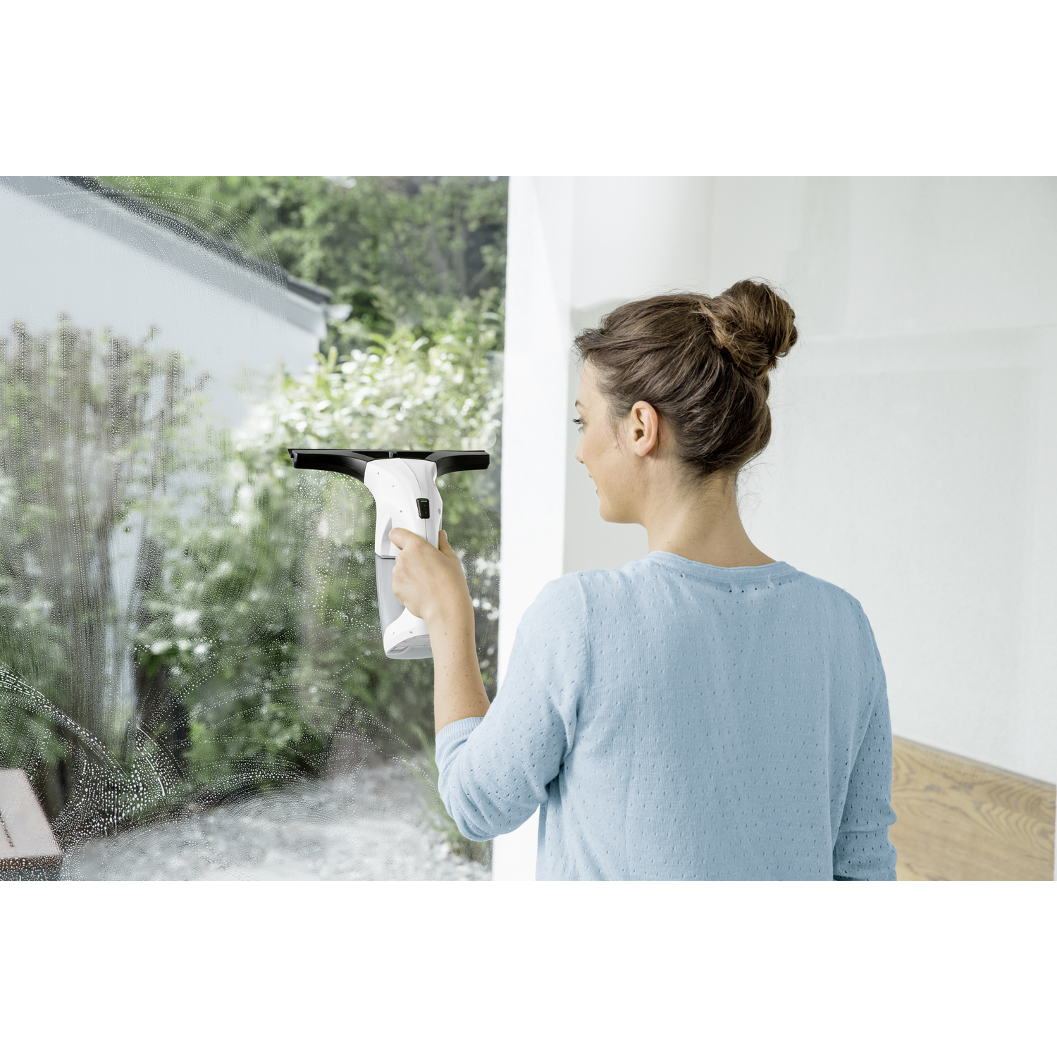 Une femme nettoie une fenêtre avec un outil de nettoyage blanc. La fenêtre est à l'extérieur. Arbres verts en arrière-plan.