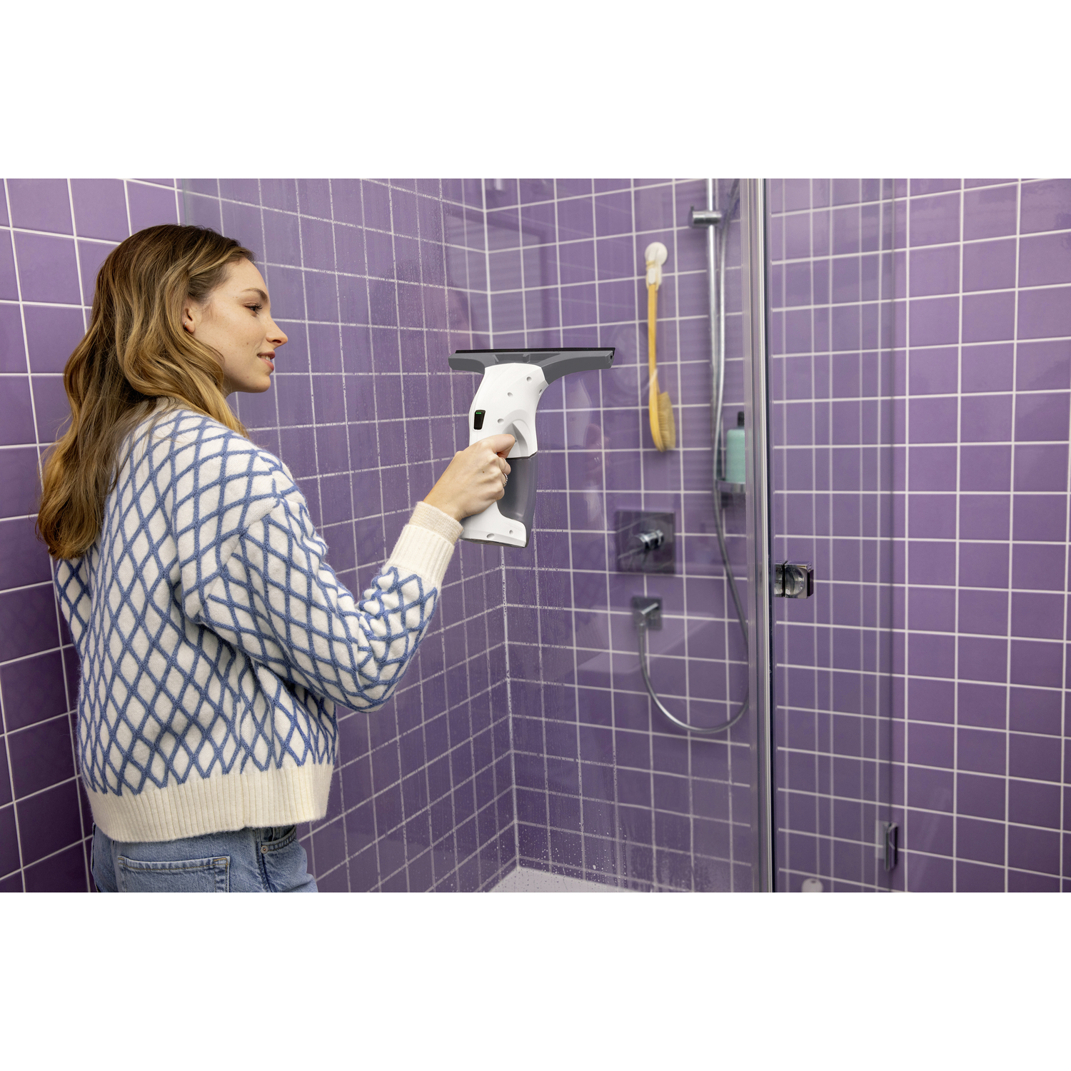 Femme nettoyant une vitre de douche. Mur carrelé violet et accessoires de douche en arrière-plan.