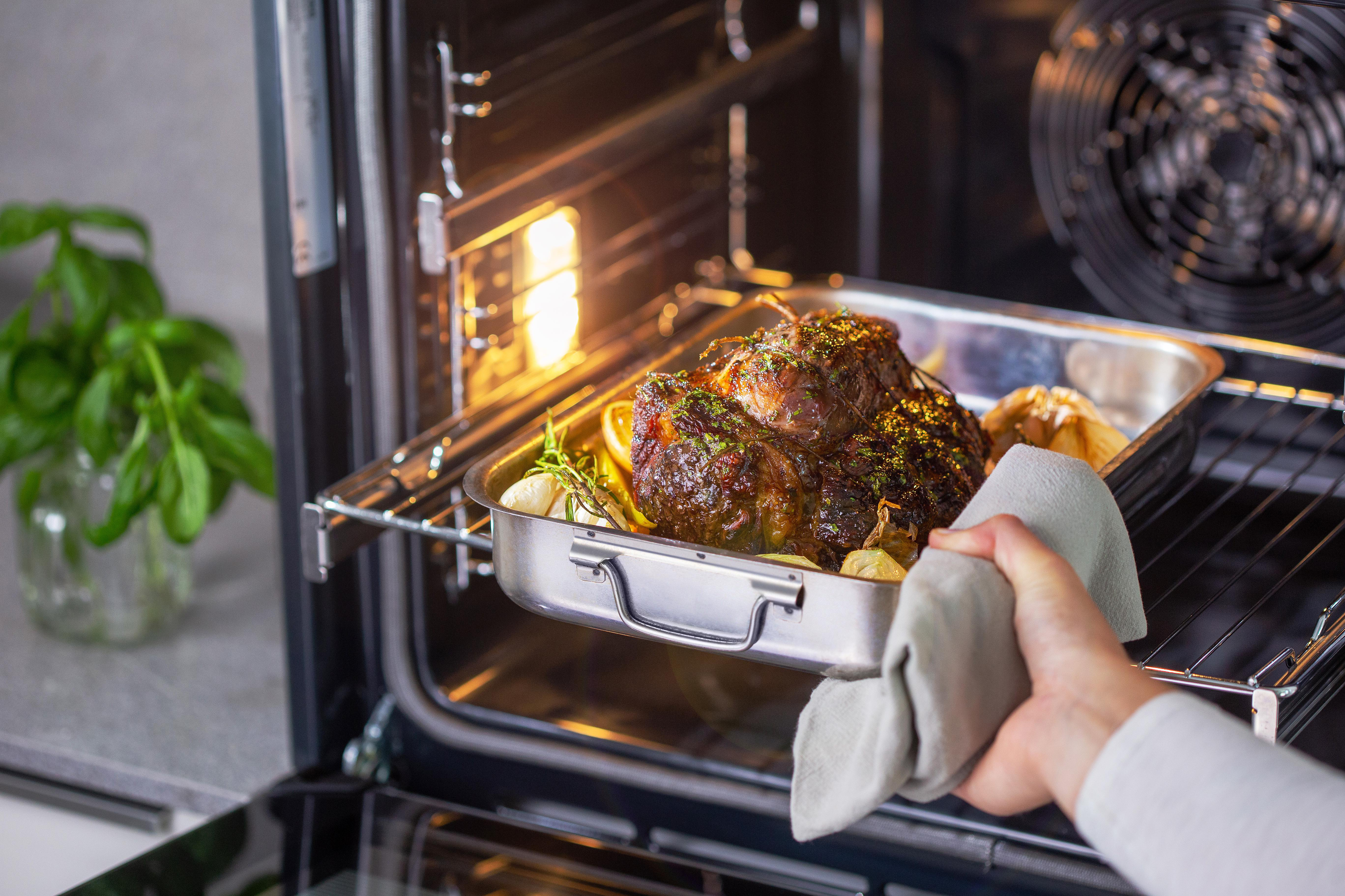 Person entnimmt eine Metallschale mit gebratenem Fleisch aus dem Ofen. Grüne Pflanze in einem Glasgefäß.