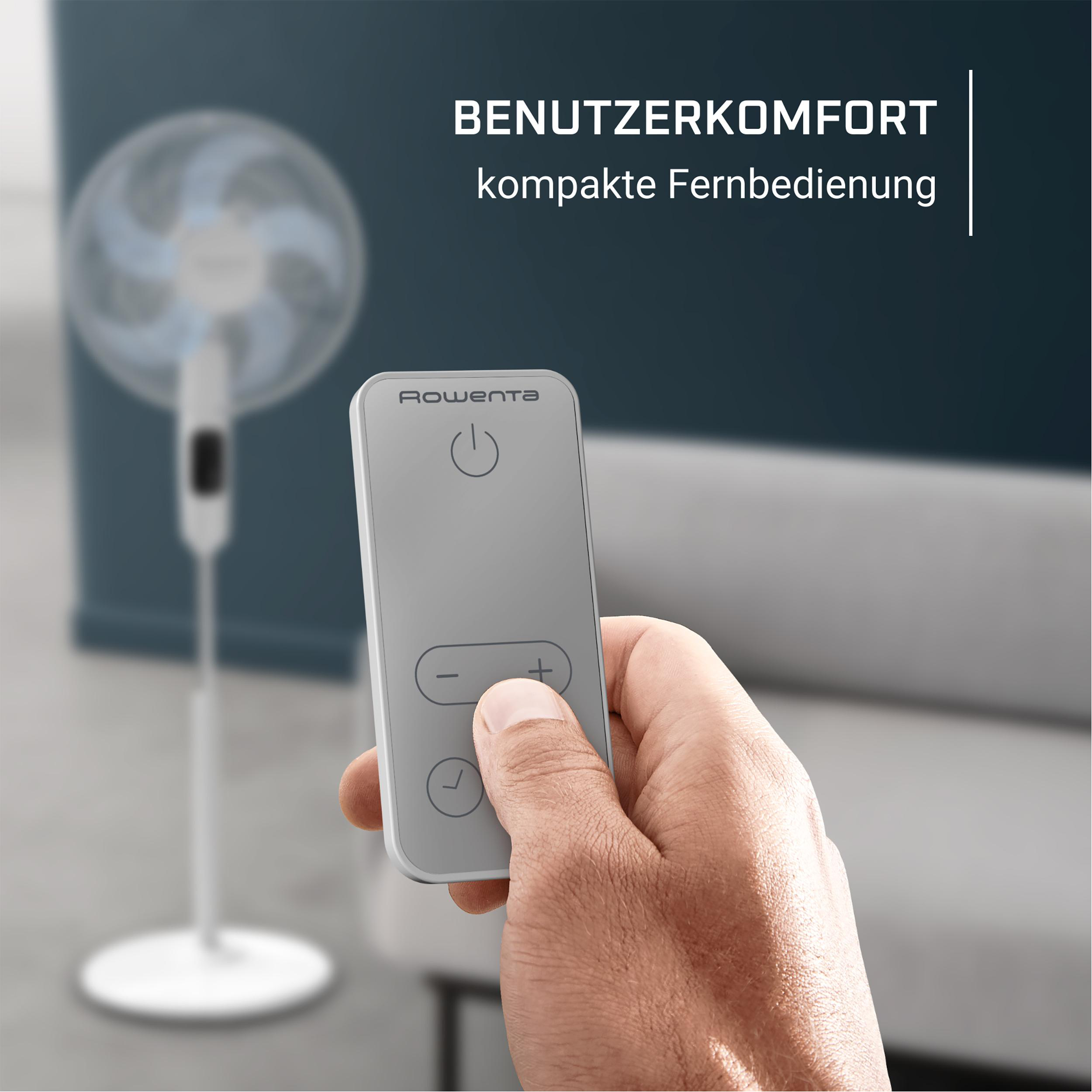 Eine Hand, die eine graue Fernbedienung mit Tasten hält, mit einem unscharfen Ventilator im Hintergrund.