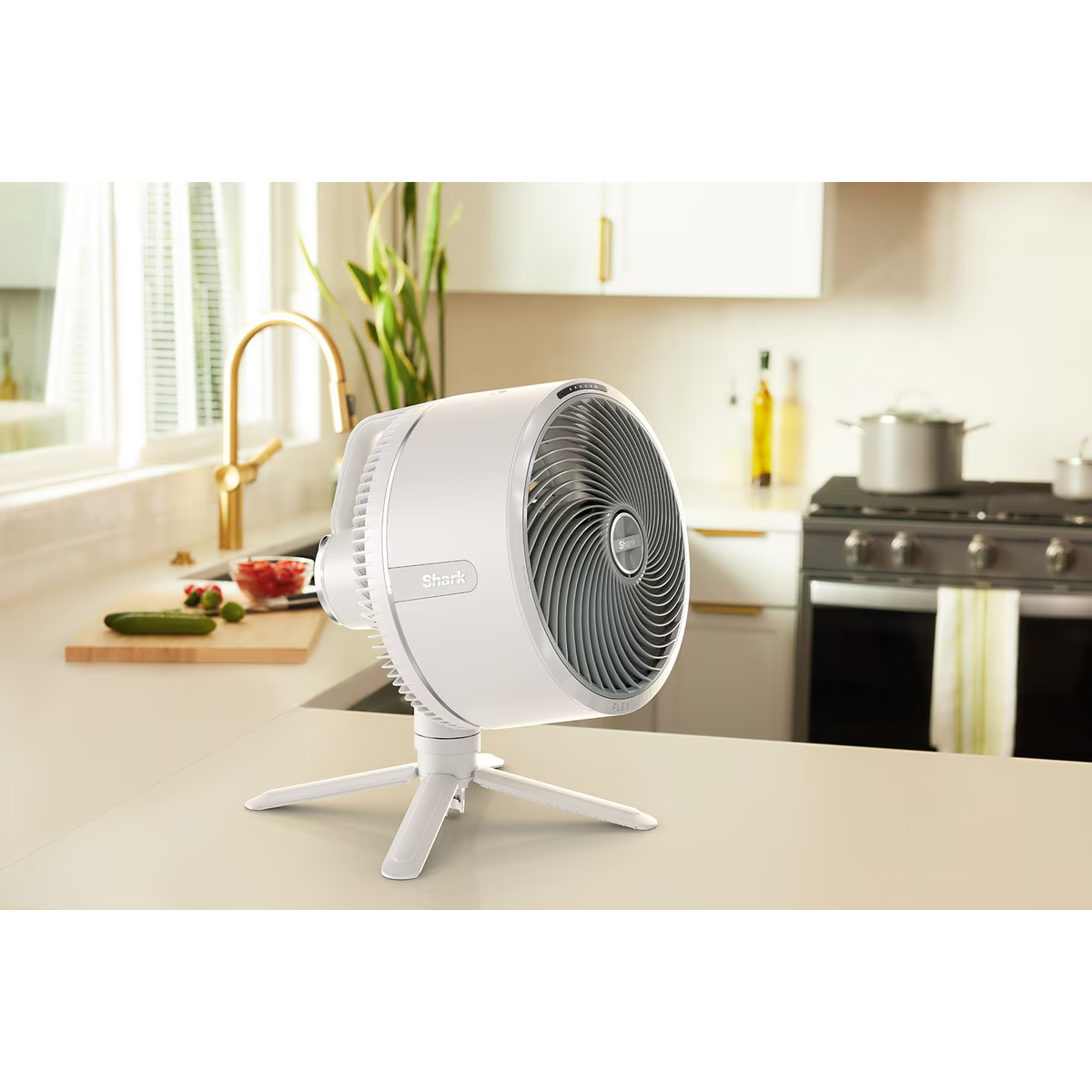 Un ventilador blanco de pie sobre un trípode blanco en una encimera de cocina, cerca de una ventana.