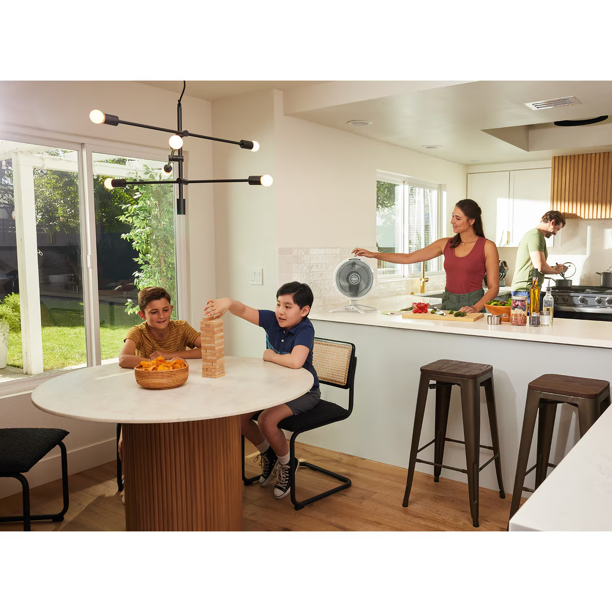 Familia en cocina jugando, mujer cocinando y ventilador en mostrador.