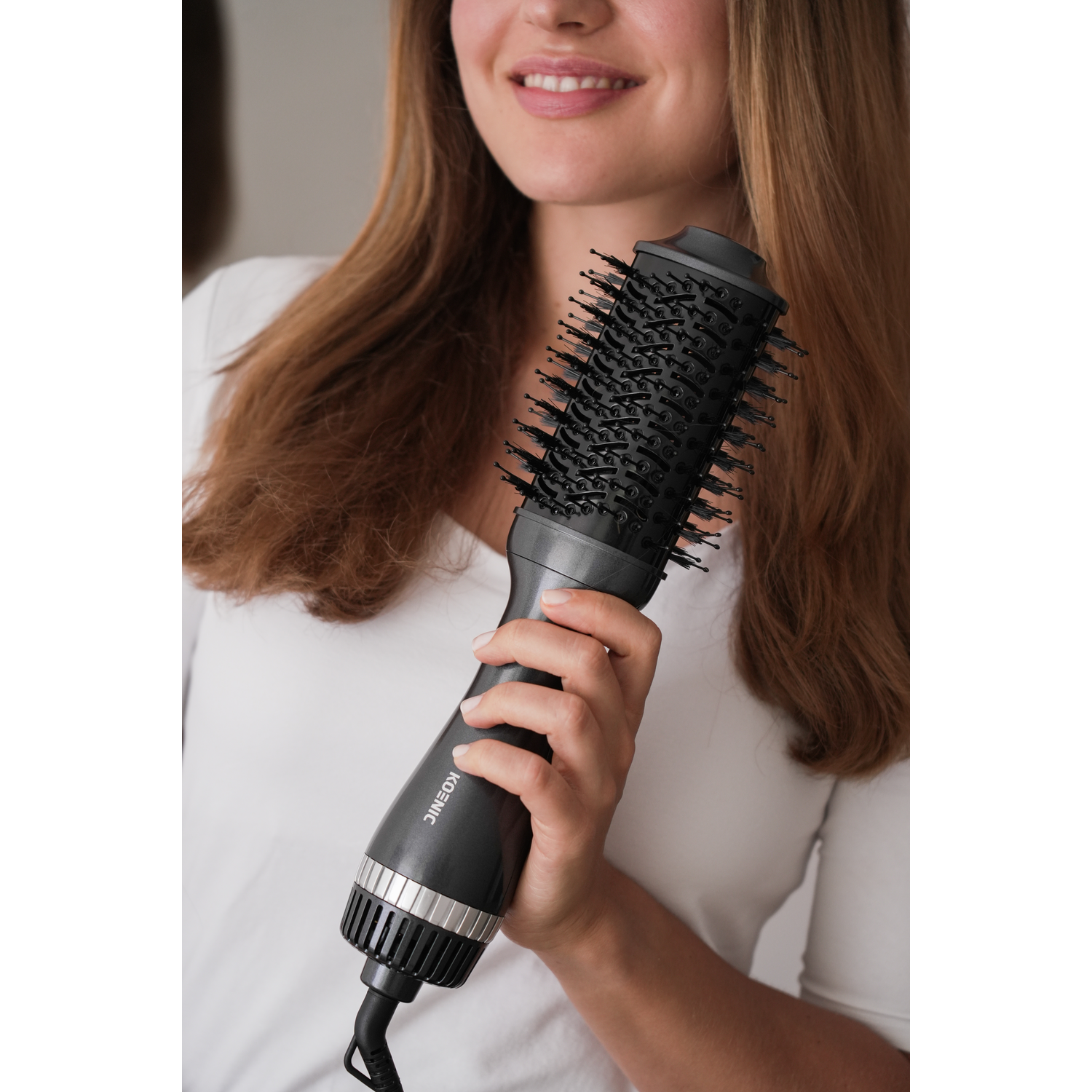 A woman holding a black hair dryer brush, smiling, with brown hair.