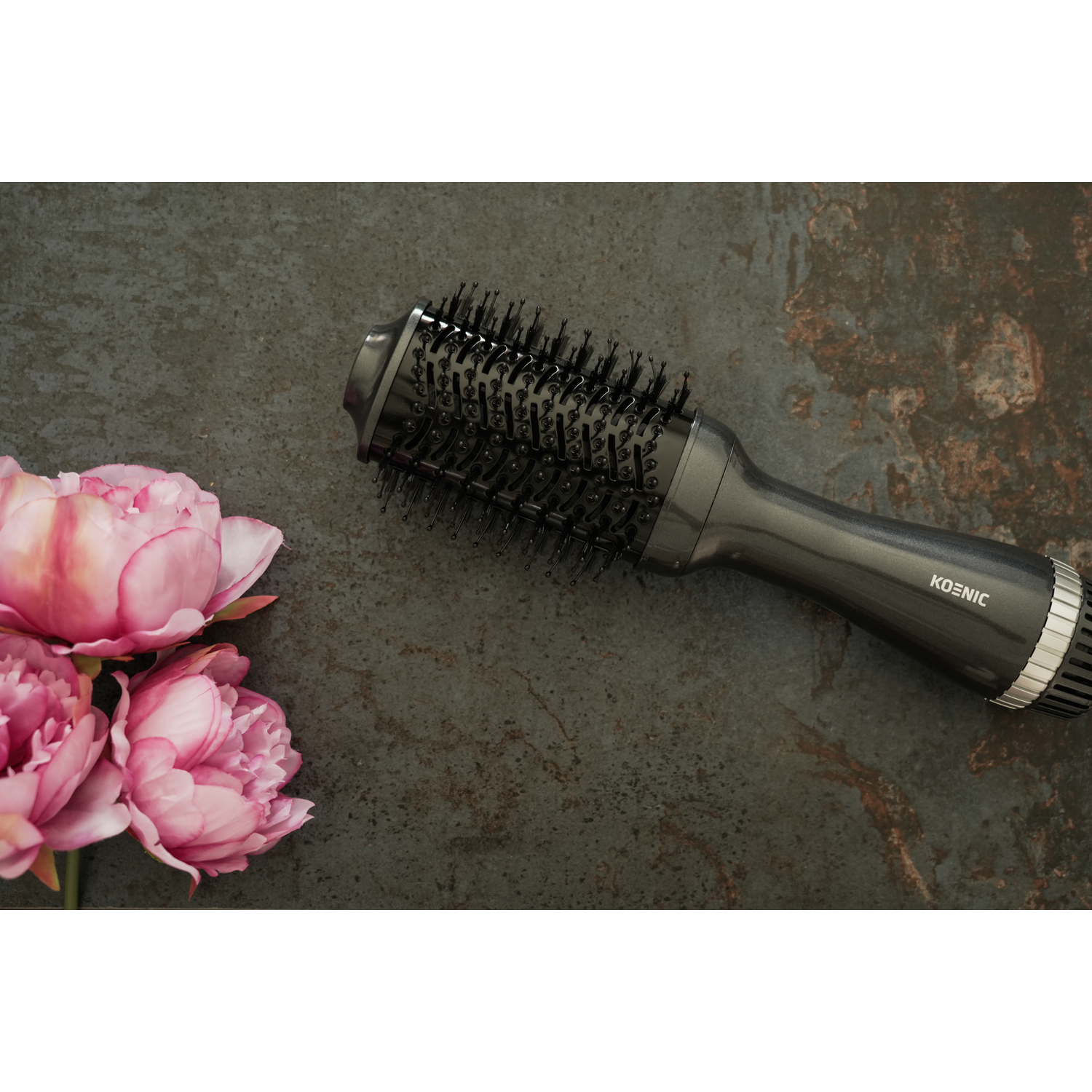 Hair styling brush next to pink flowers on a textured dark surface.