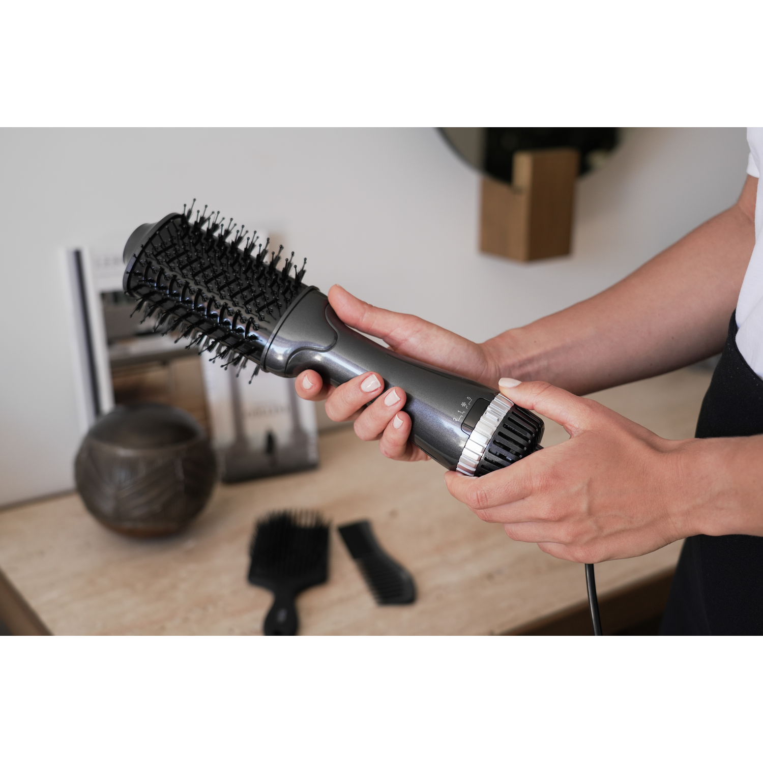 A woman holding a gray hair dryer brush, with a black brush and comb in front.