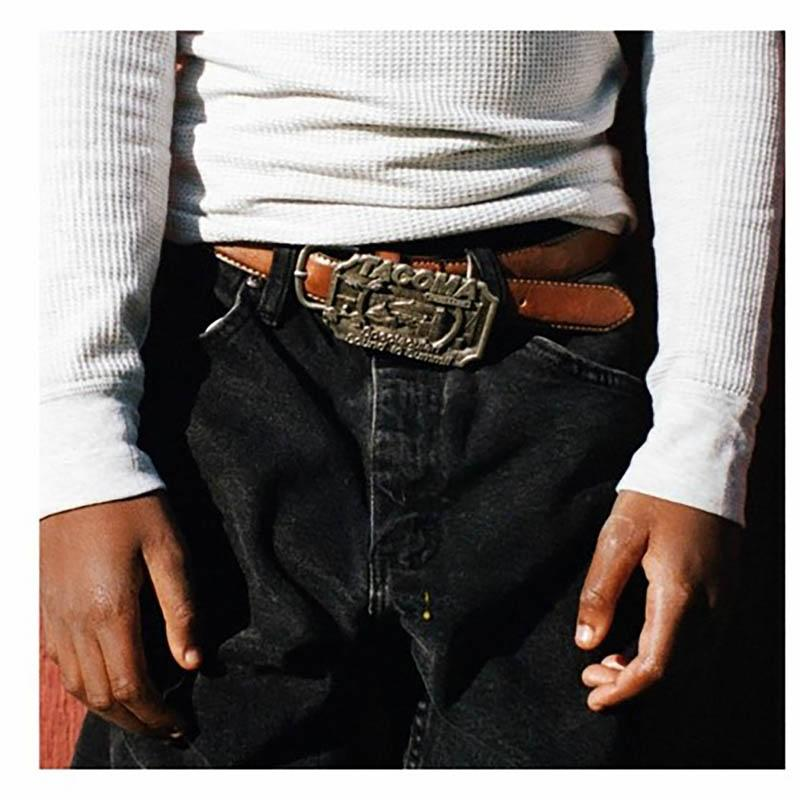 Eine Person trägt Jeans, ein weißes Hemd und einen braunen Gürtel mit Schnalle und Logo.