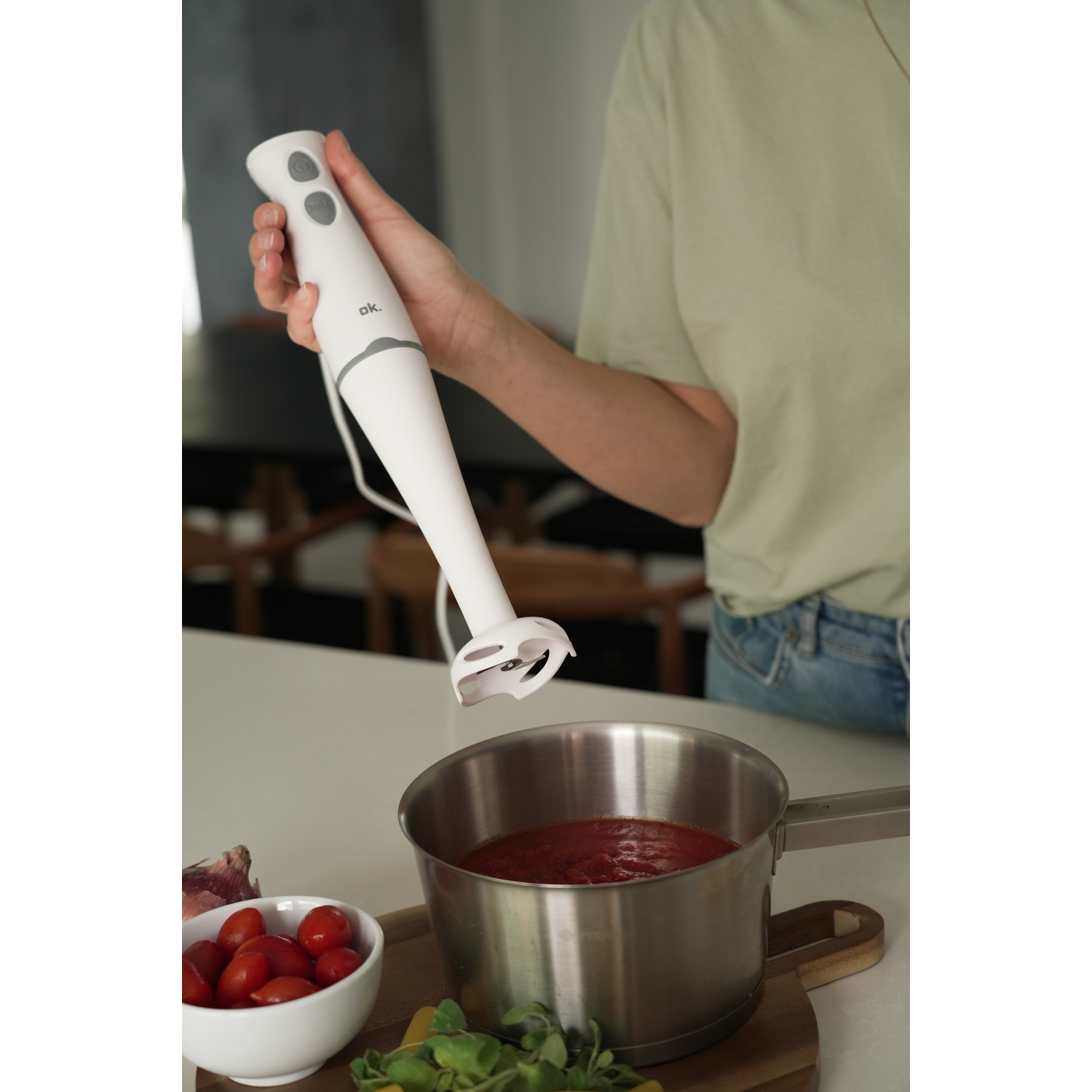 A person using a white hand blender over a pot of tomato sauce. A bowl of tomatoes is nearby.