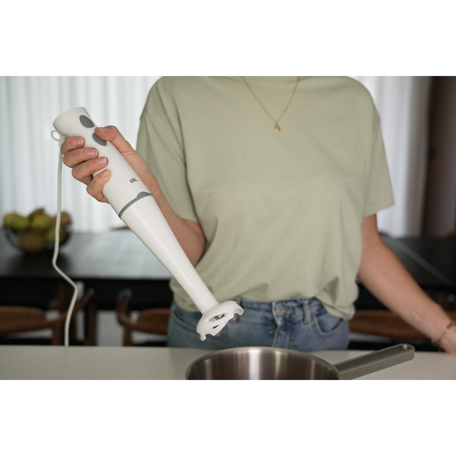 A person holding a white hand blender over a metal pot on a white countertop.