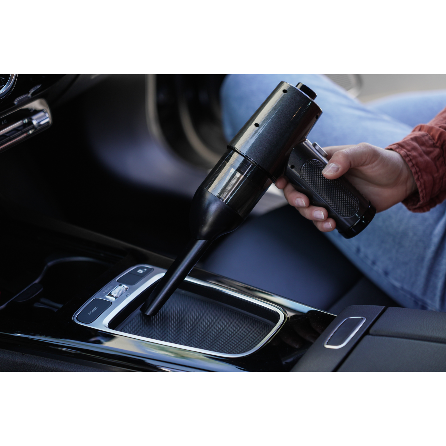 A black handheld vacuum cleaner is used inside a car, near the gear stick.