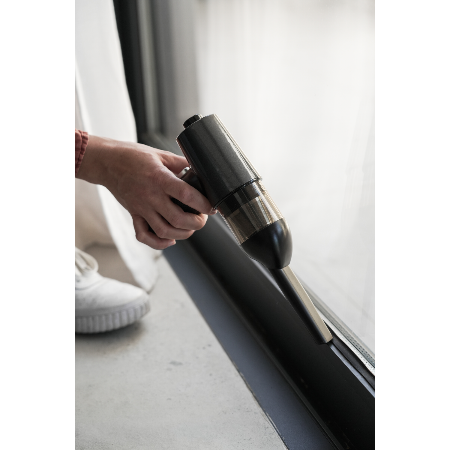Person's hand using a vacuum cleaner to clean a window frame. White shoe, grey floor.