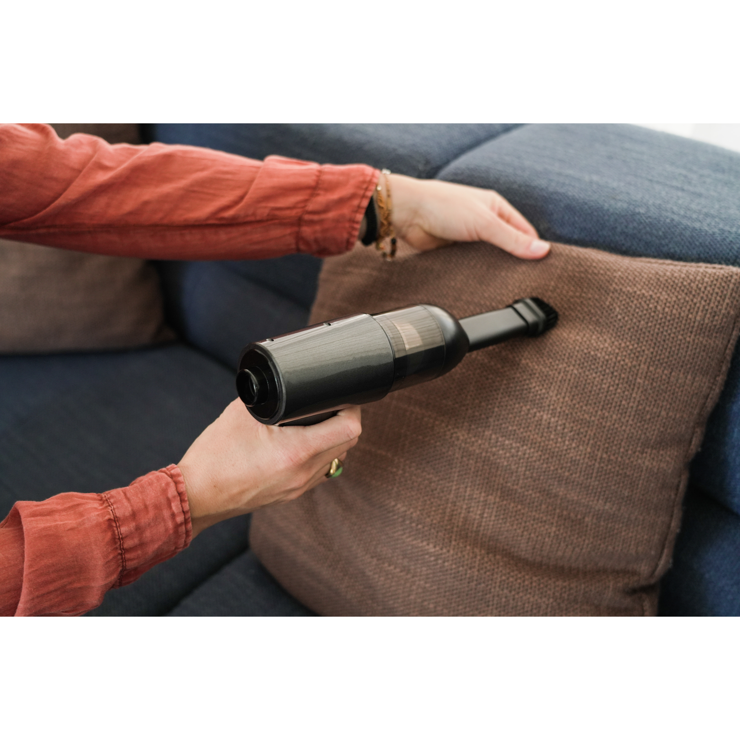 A person using a black handheld vacuum cleaner to clean a brown cushion on a blue sofa.