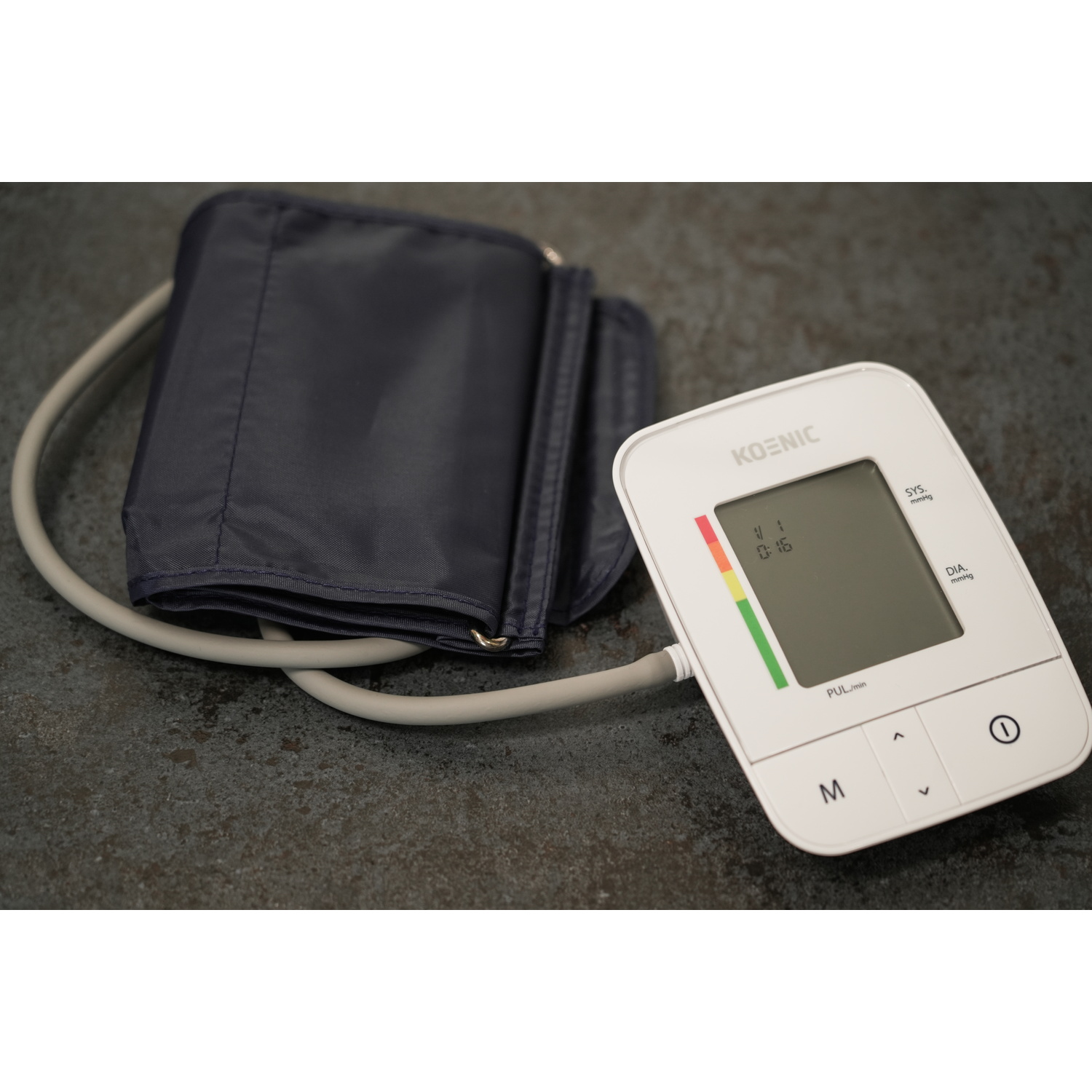 Blood pressure monitor with cuff. White display shows readings. Gray cord and a dark surface.