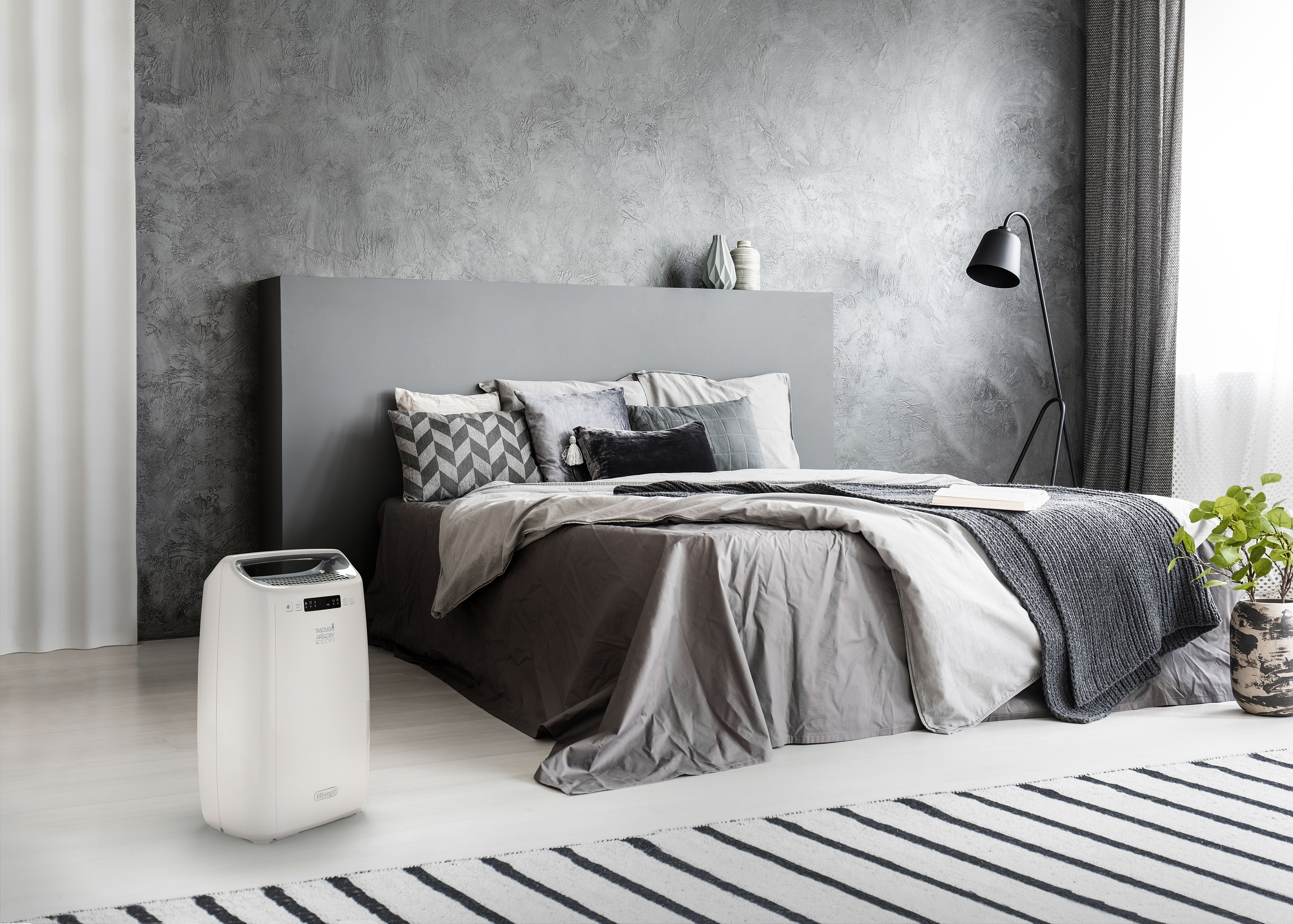 Un dormitorio con paredes grises, cama y un purificador de aire DeLonghi blanco en el suelo.