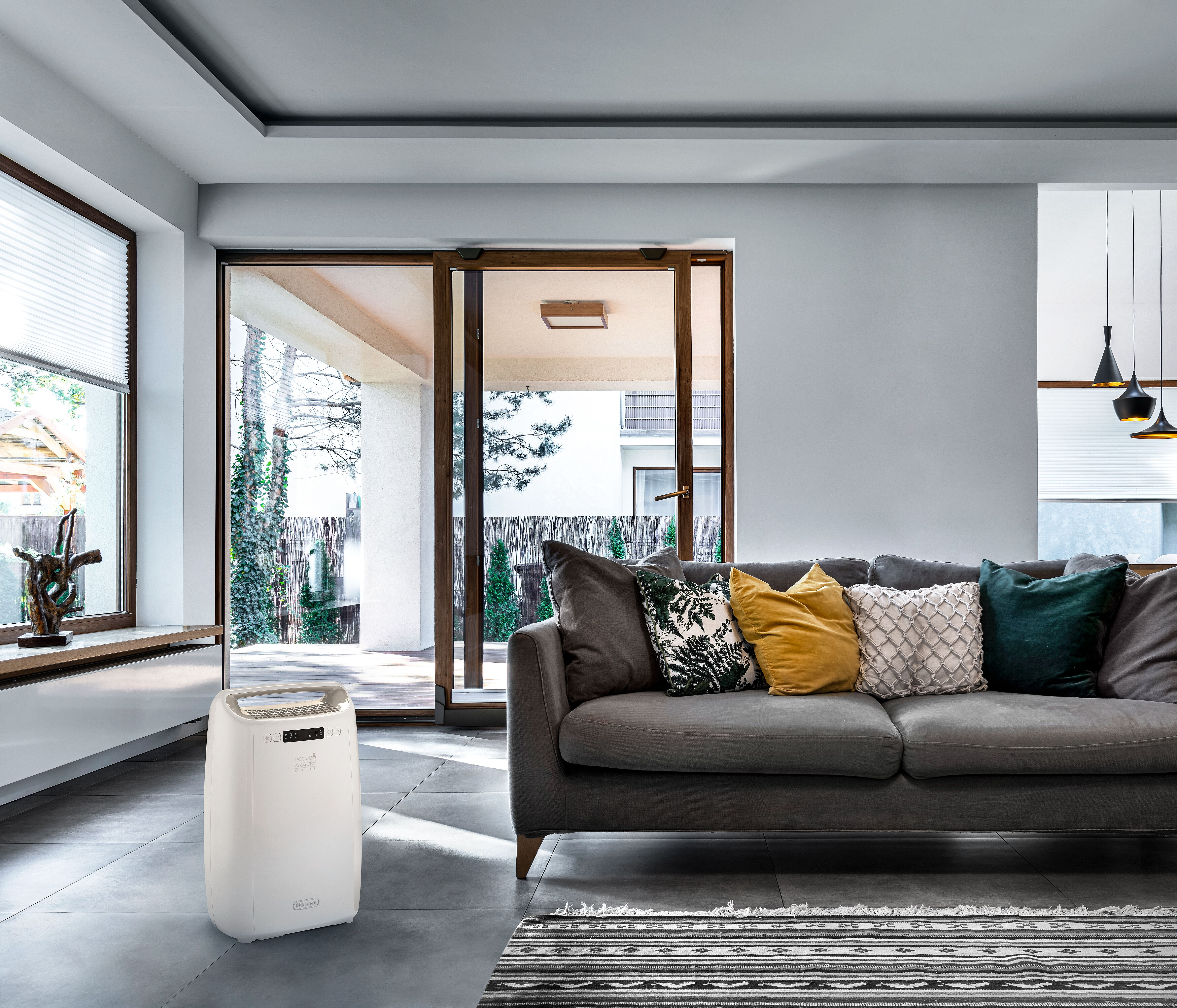 Un deshumidificador blanco en una sala de estar con sofá, ventanas y vista al exterior.
