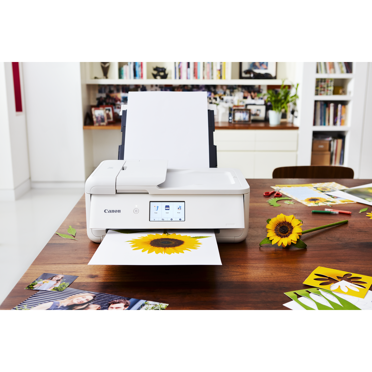 Printer printing a sunflower picture, with other photos and a flower on a table.