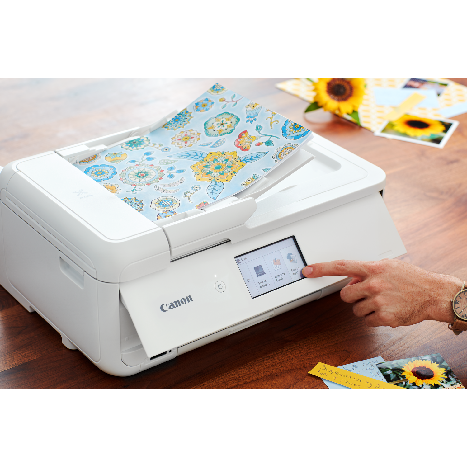A white printer with a floral design paper inside, a hand presses its screen. Sunflower and printouts are on the table.