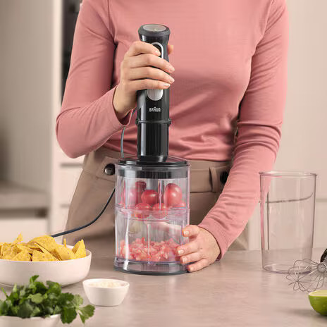 Blender z czerwonymi pomidorami, kobieta go używa, miska z chipsami i kolendrą.