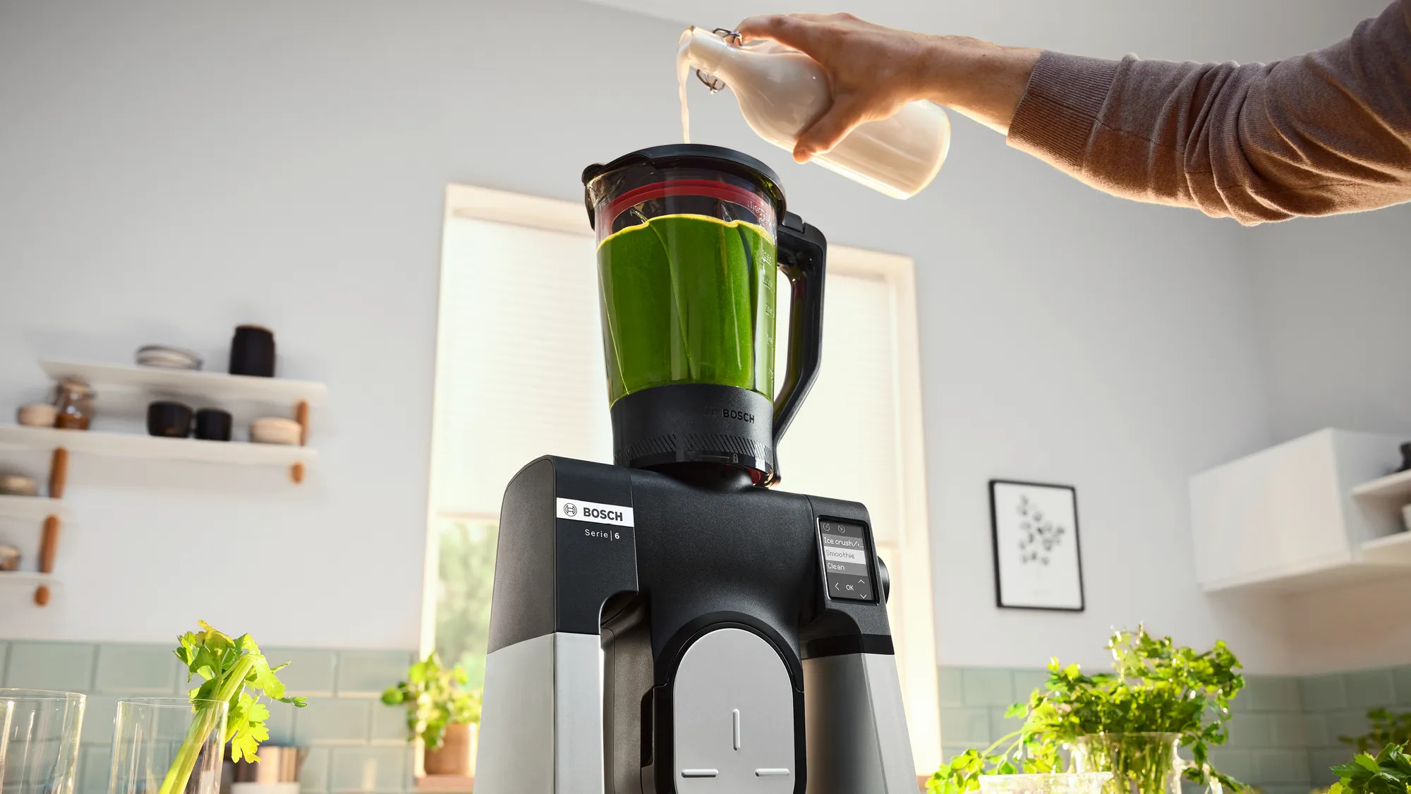 Osoba wlewa płyn do zielonego blendera. Seler i inne składniki na pierwszym planie.