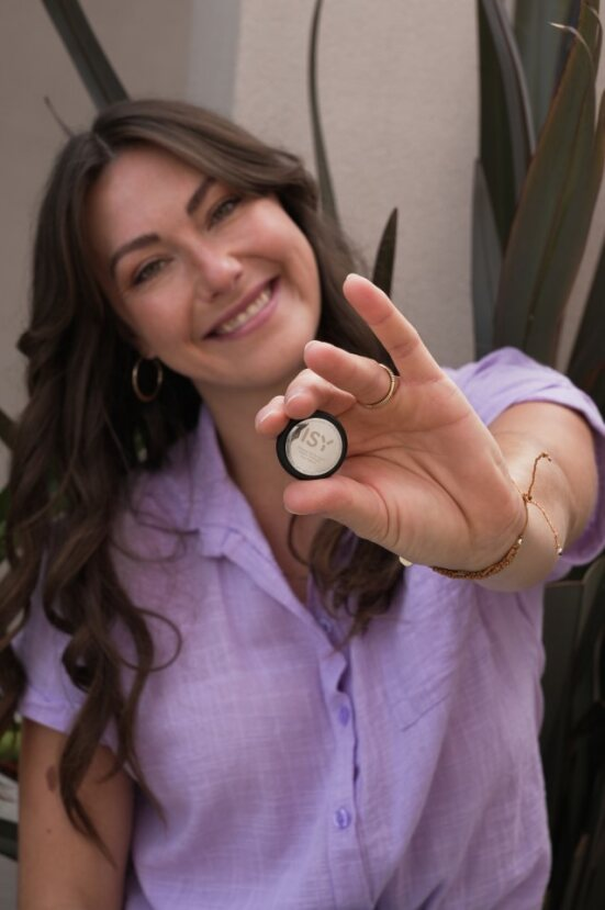 Donna che tiene in mano un piccolo oggetto rotondo, sorridente. Fogliame verde sullo sfondo.