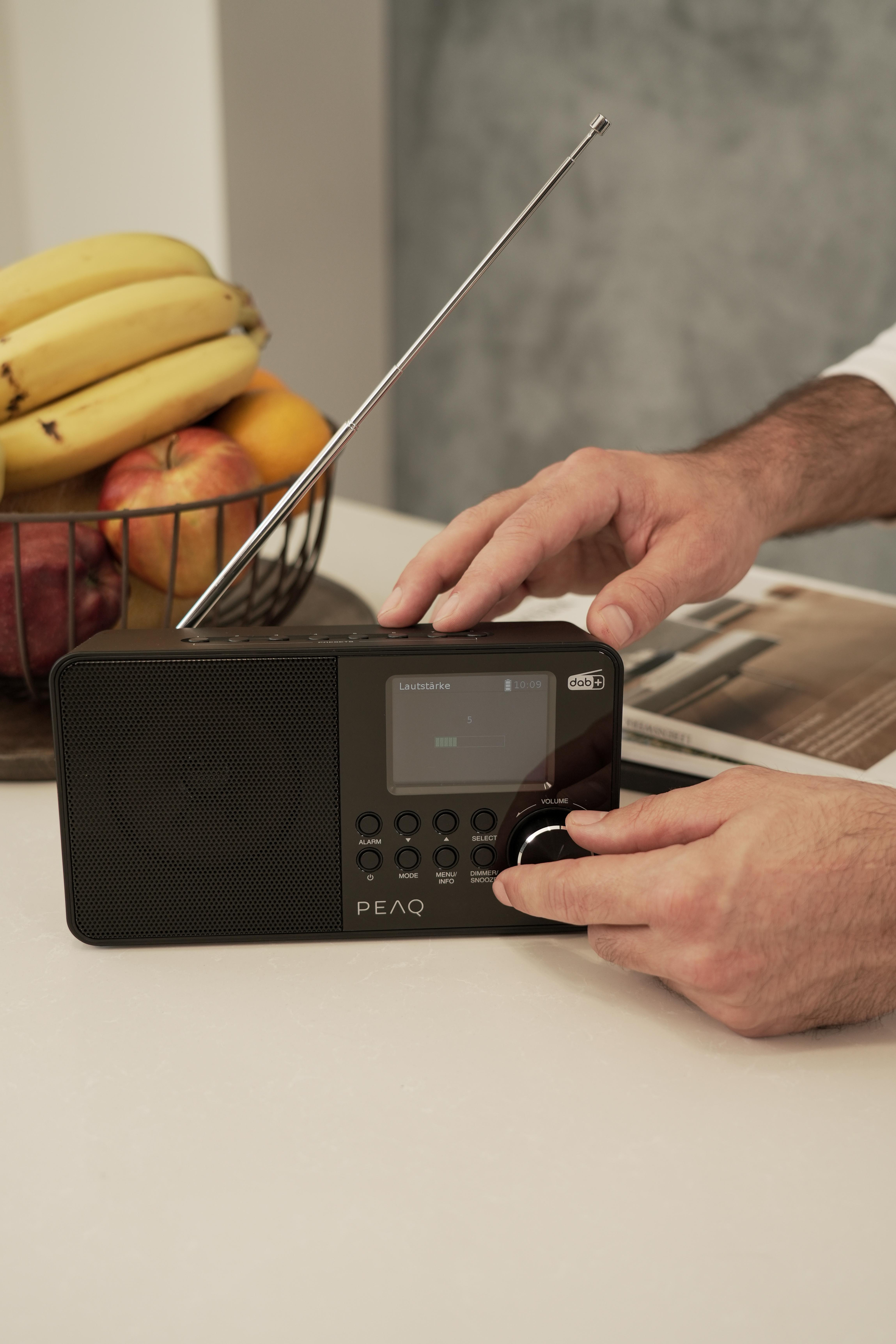 Ein Mann stellt den Lautstärkeregler an einem schwarzen Radio ein, mit Obst und einer Zeitschrift in der Nähe.