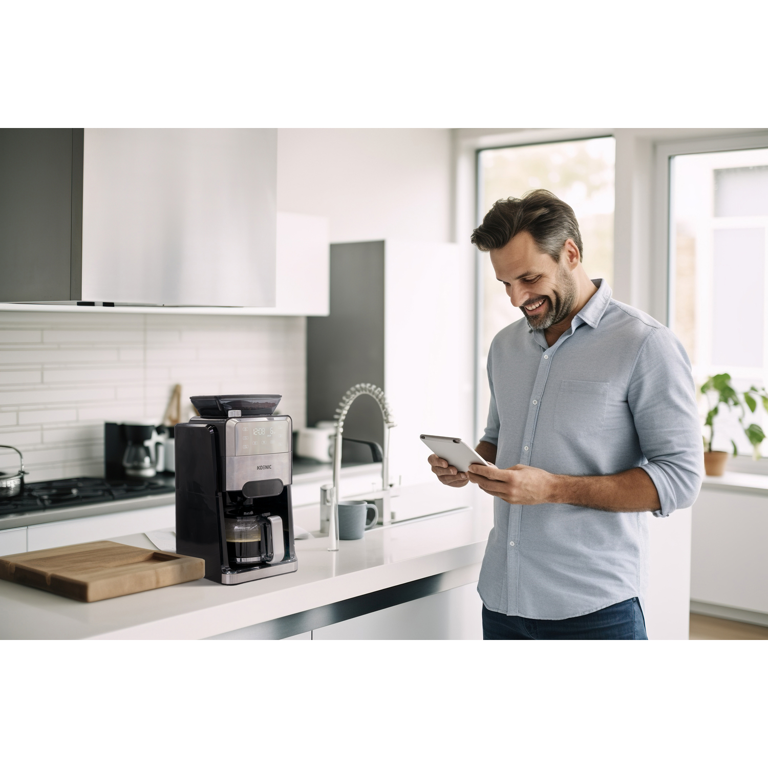 Man in a kitchen using a tablet next to a coffee maker. Kitchen is modern with white cabinets.
