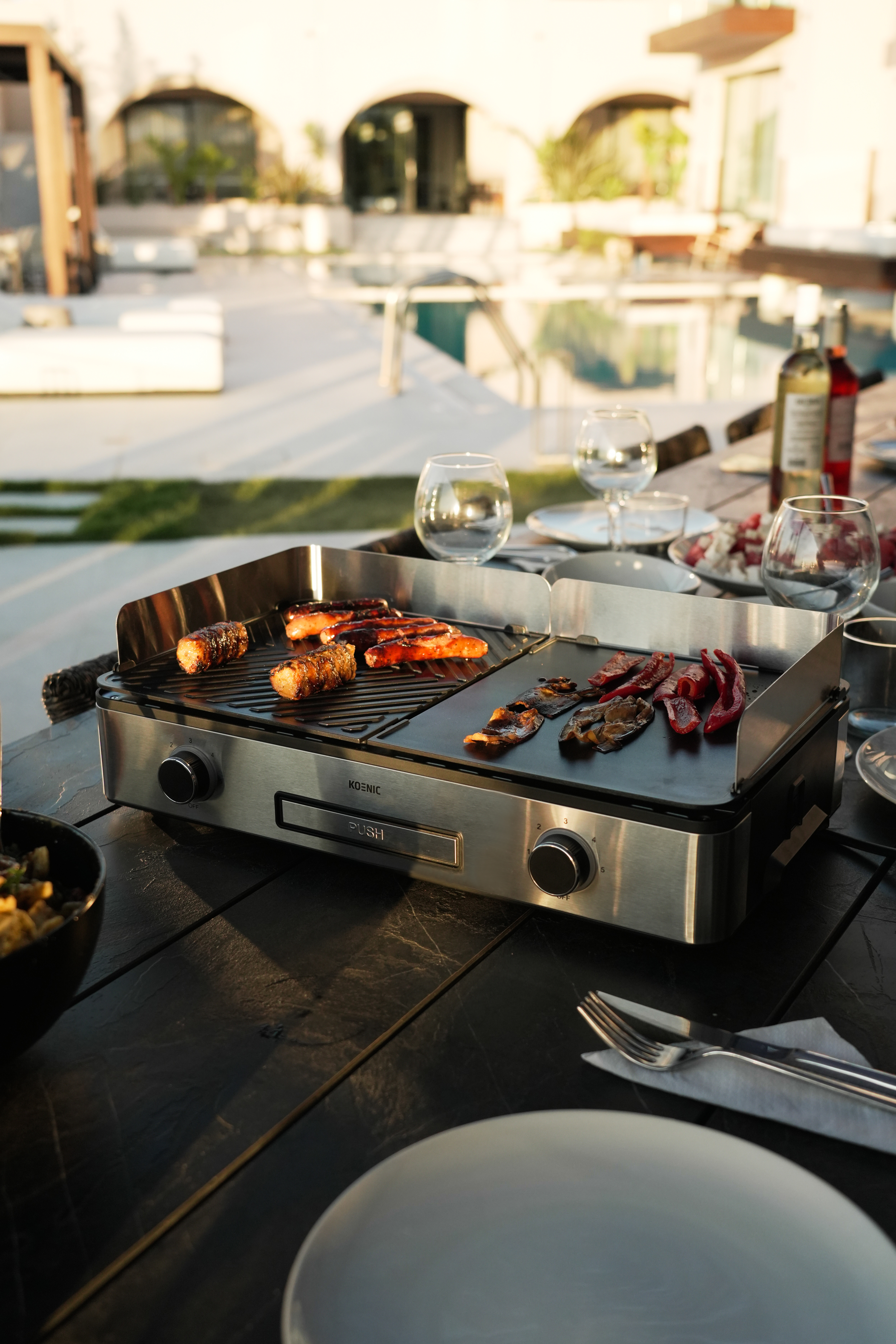 Grill mit Würstchen auf dem Tisch, Gläsern und Pool im Hintergrund.