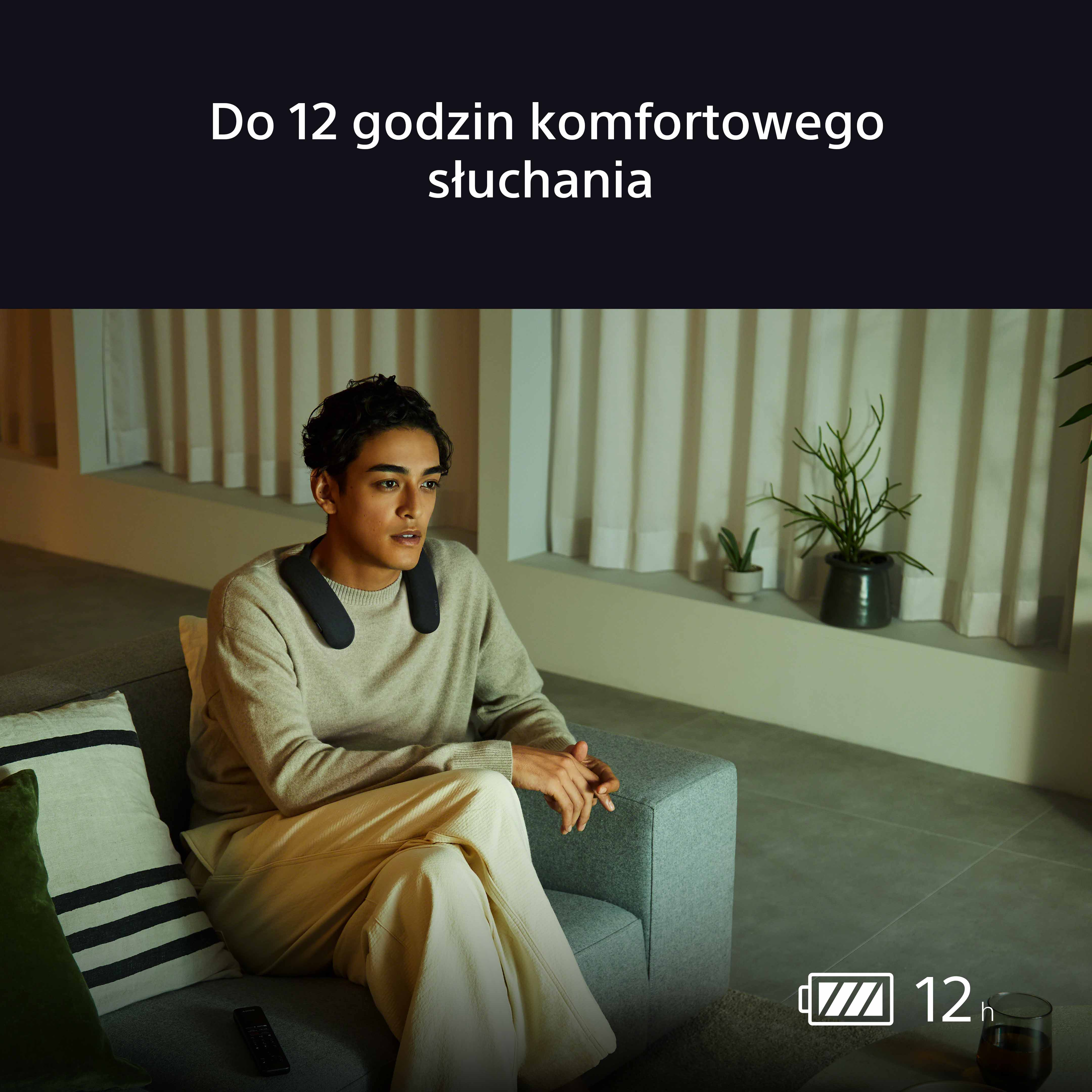 Mężczyzna noszący głośnik na szyi siedzi na kanapie, z ikoną baterii.