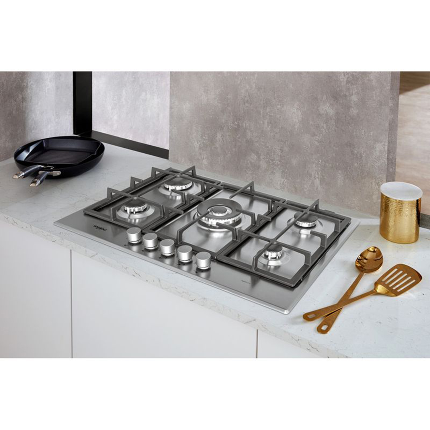 Piano cottura a gas in acciaio inox con cinque fuochi, su piano bianco, con utensili da cucina.