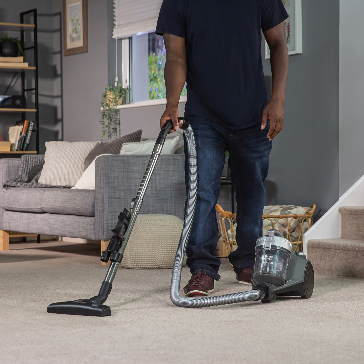 Mann, der mit einem Staubsauger Teppich saugt. Graues Sofa und Treppe im Hintergrund.