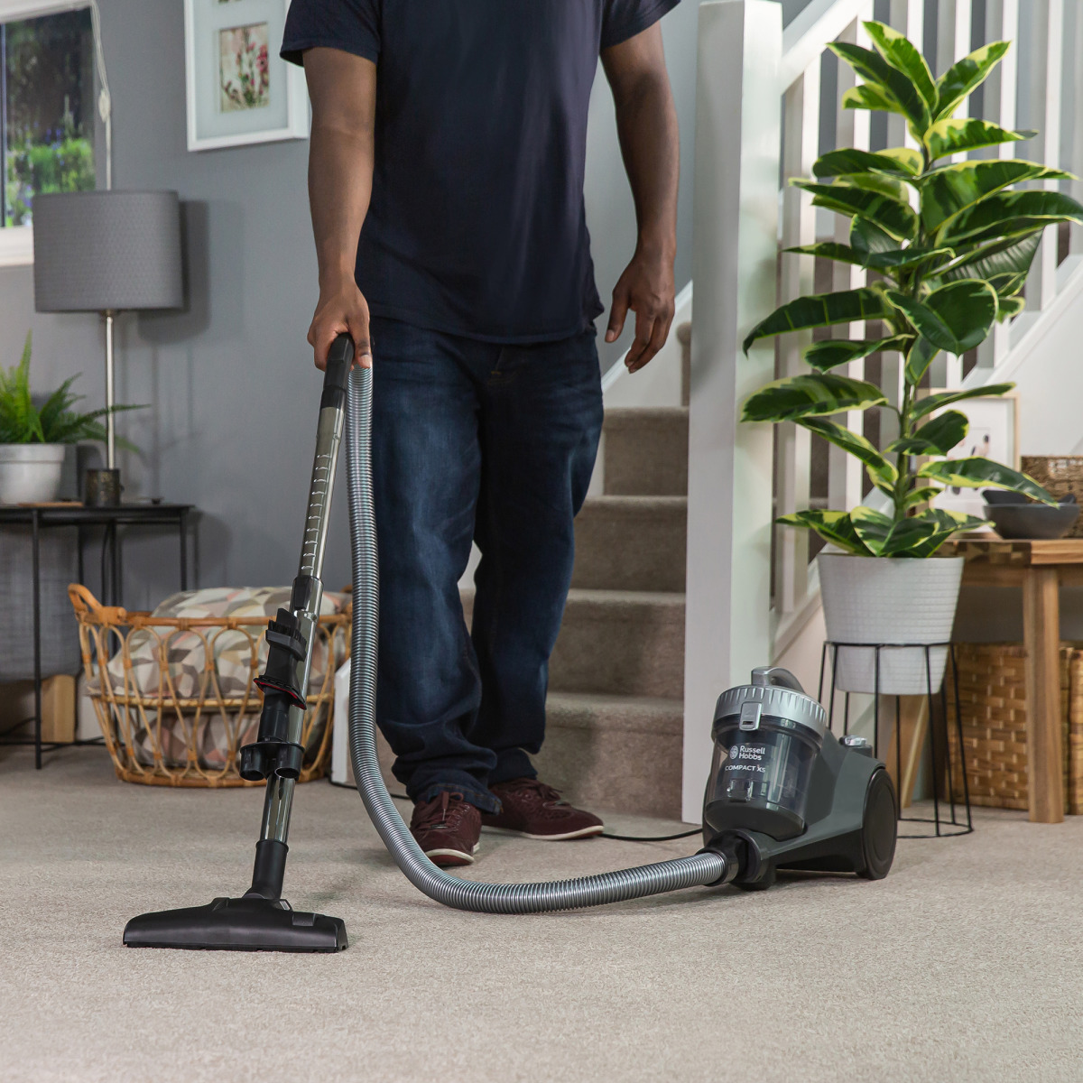 Mann saugt beigen Teppich. Staubsauger ist grau und schwarz. Treppe und Pflanze im Hintergrund.