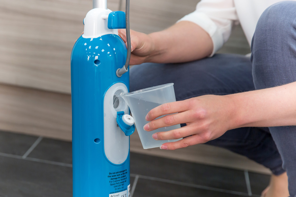 Persona che versa acqua nel mocio a vapore blu e bianco, vicino a una cucina.
